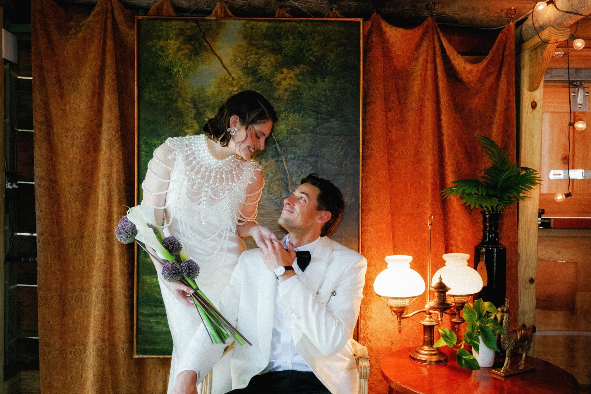 A woman in a white dress holding flowers stands smiling, gazing at a seated man in a white tuxedo. At their Caya Azul Asheville wedding, they are framed by orange curtains, green plants, a classic lamp, and an elegant painting.