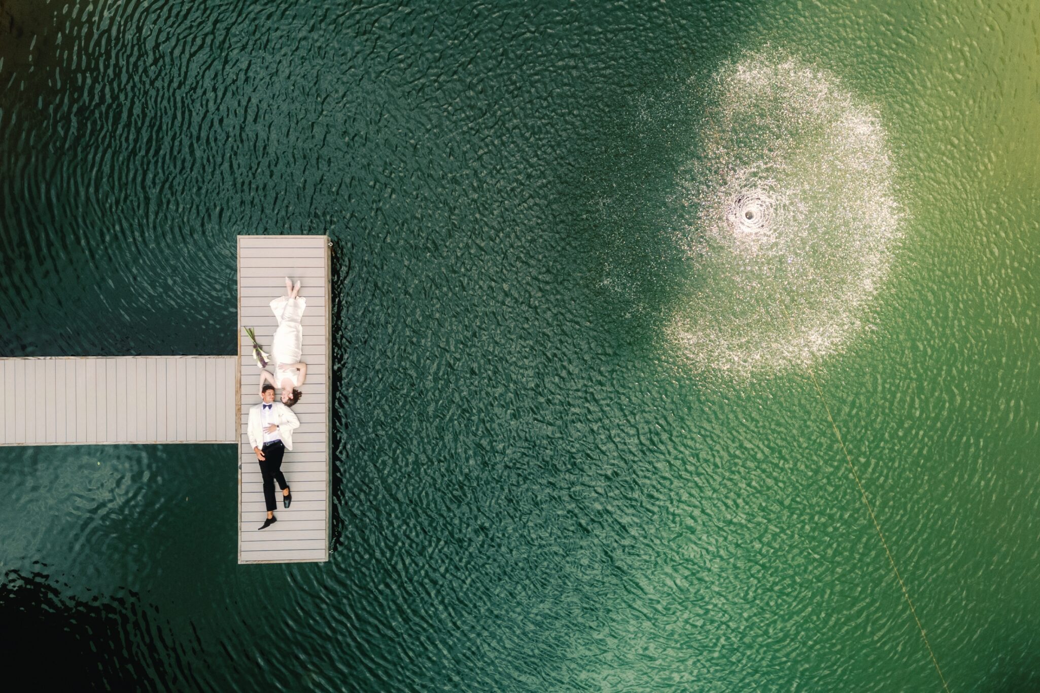 A couple in wedding attire lies on a dock extending into a green lake, near a circular water fountain. The aerial view highlights gentle ripples around the dock and the bright fountain on the right.