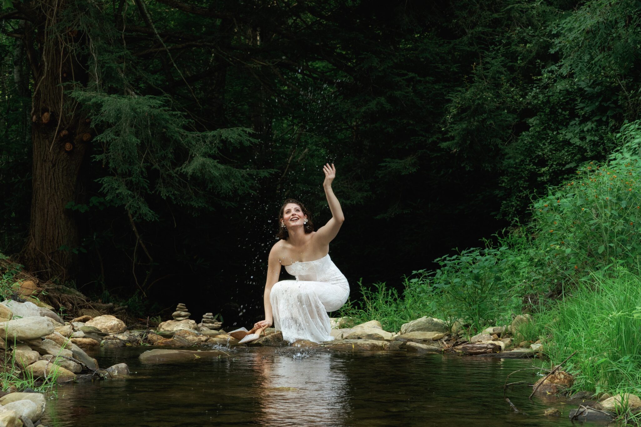 A woman in a white dress sits on rocks by a stream in a lush green forest, smiling joyfully while splashing water with one hand.