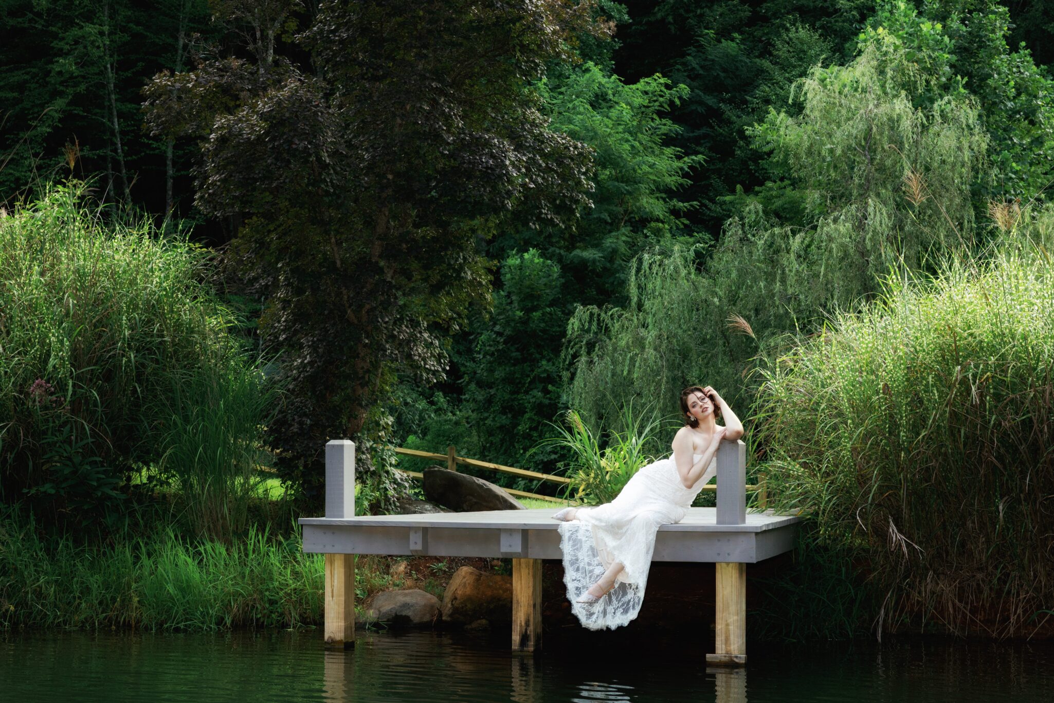 A woman in a white dress reclines on a wooden dock over the water, surrounded by lush green trees and tall grasses at a serene Caya Azul Asheville wedding.