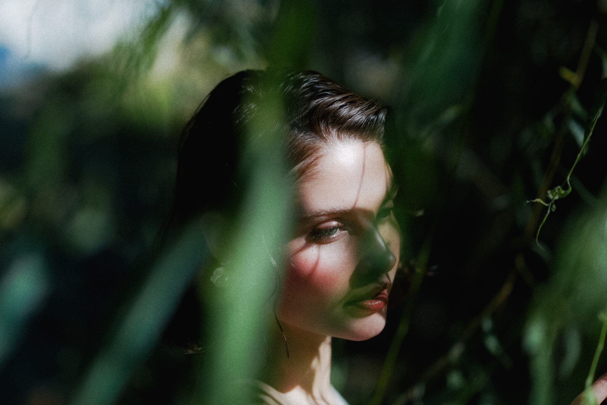 A woman with fair skin and dark hair stands among greenery, partially obscured by blurred leaves, with sunlight casting shadows on her face and a contemplative expression.