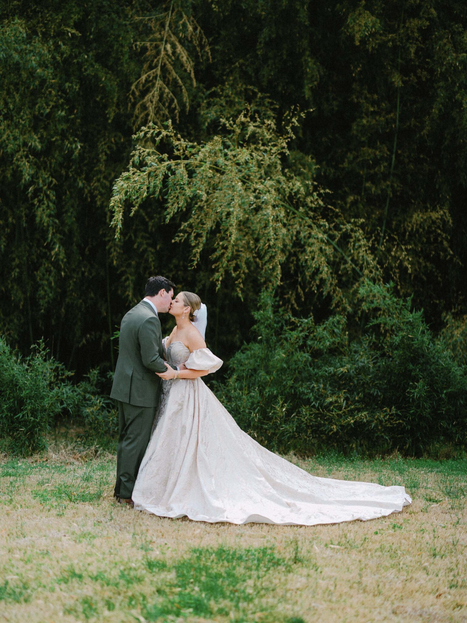 A bride and groom share a kiss outdoors, surrounded by greenery. The bride wears a flowing white gown with a long train, while the groom is dressed in a green suit. They stand on grass under tall trees.