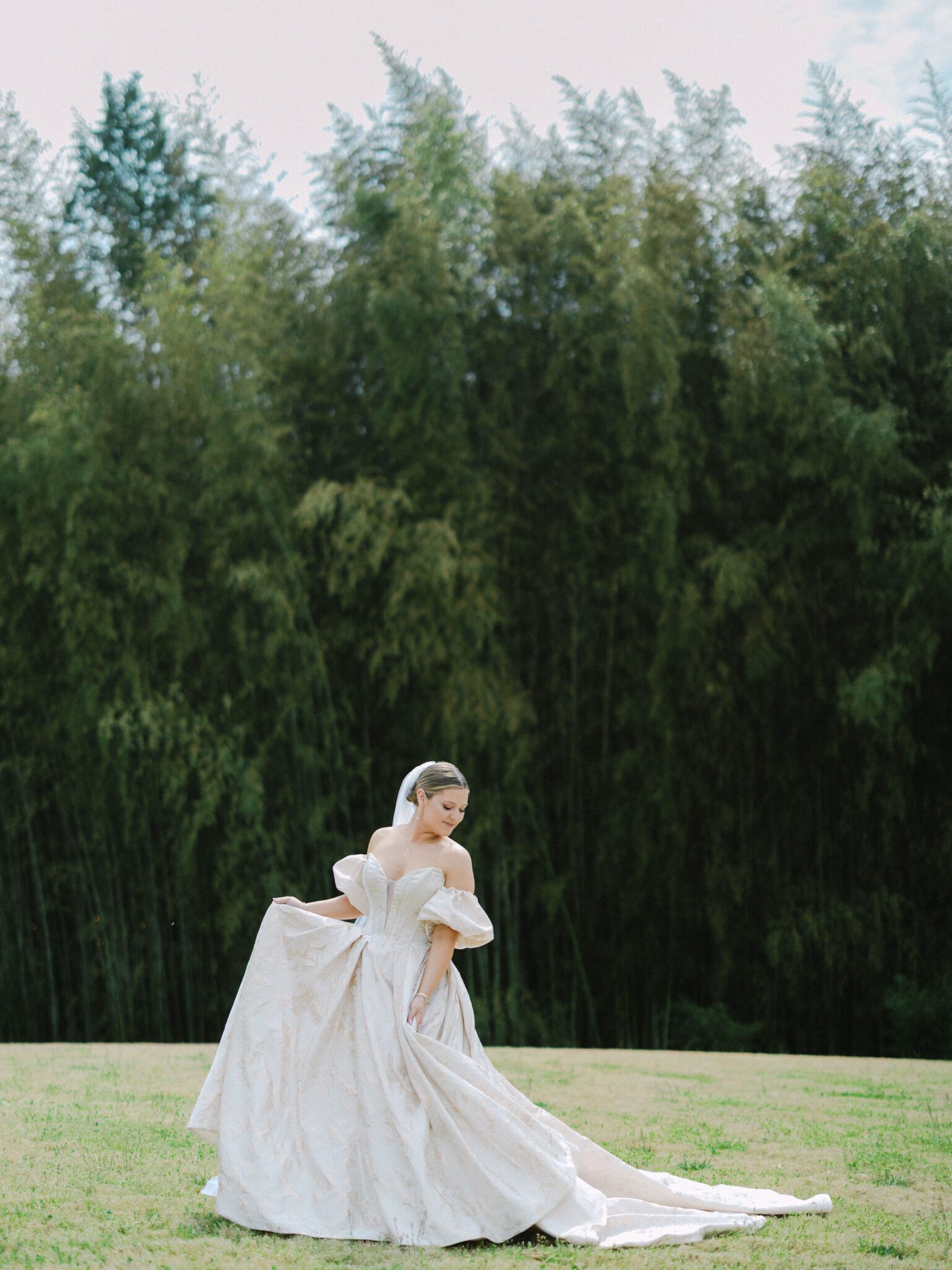 A woman in an elegant white wedding gown and veil stands on green grass at Camelot Meadows, holding her dress, with a backdrop of tall, dense trees under a cloudy sky—captured on 35mm film near Charlotte, NC.
