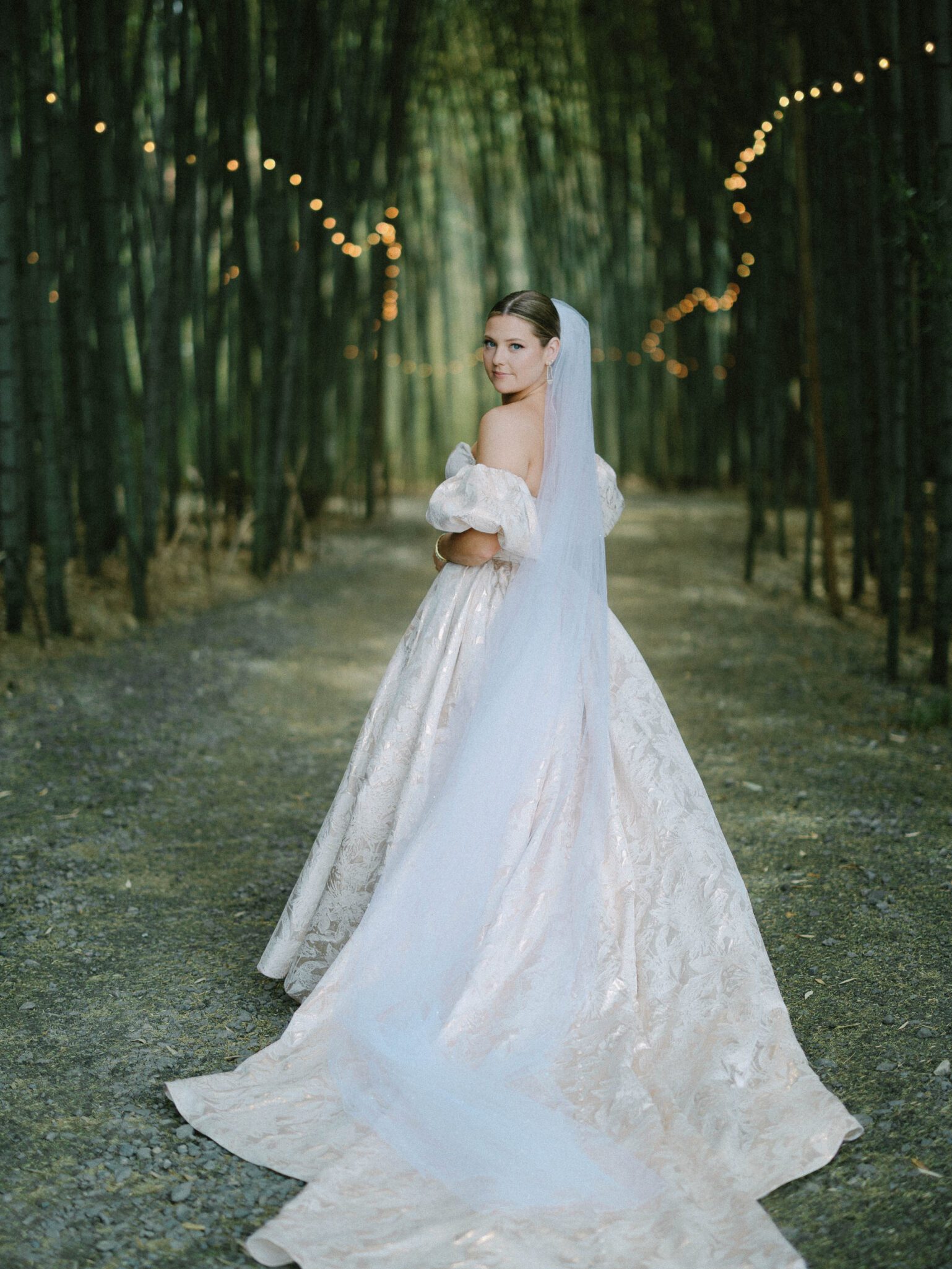 A bride in an off-the-shoulder, textured gown with a long veil stands on a forest path lined with string lights, looking over her shoulder toward the camera.