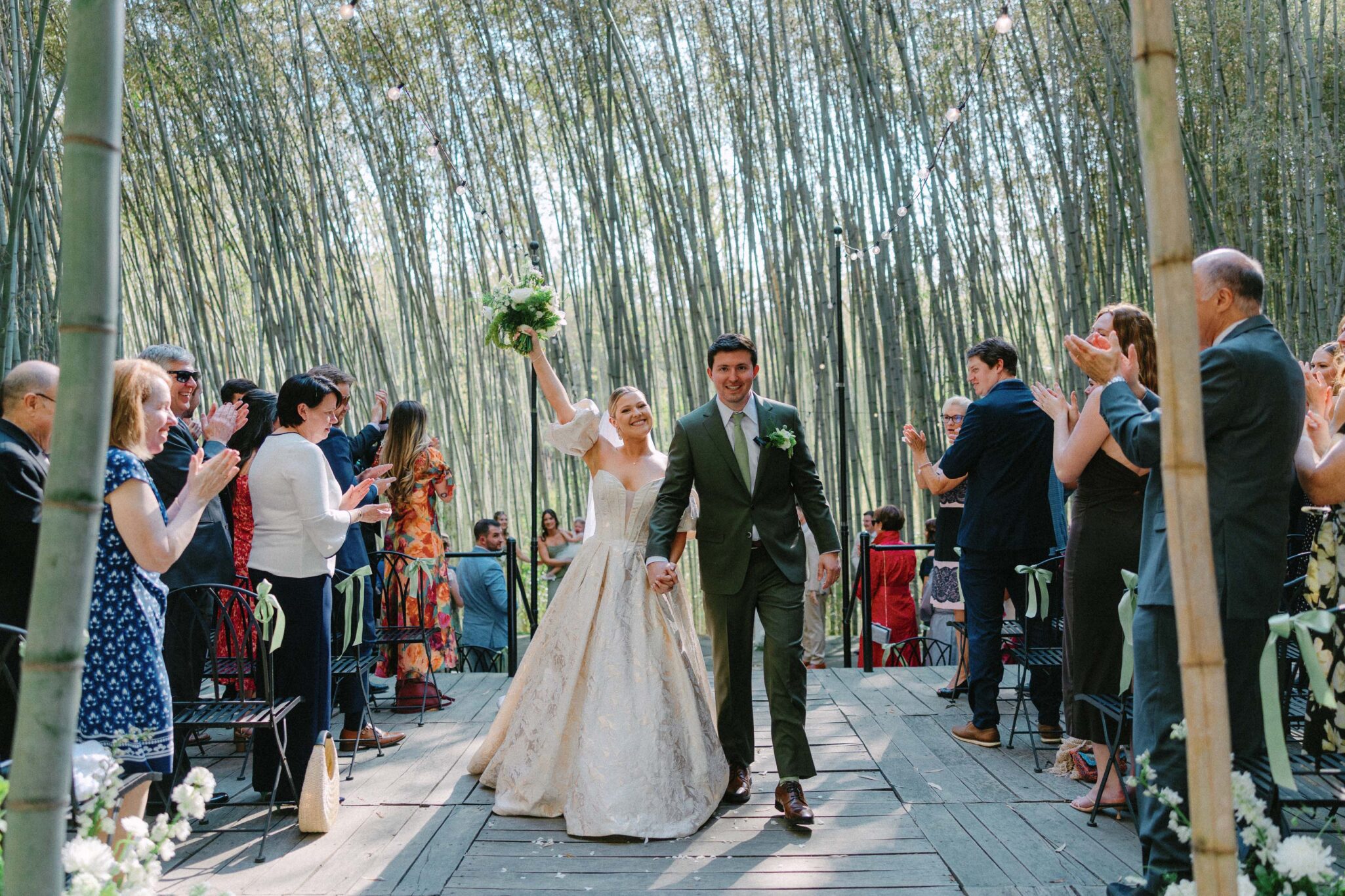 At Camelot Meadows in Charlotte, NC, a bride and groom walk down a bamboo-lined outdoor aisle as guests cheer. Captured on 35mm film, the joyful couple beams with celebration, the bride lifting her bouquet while holding hands with her groom.
