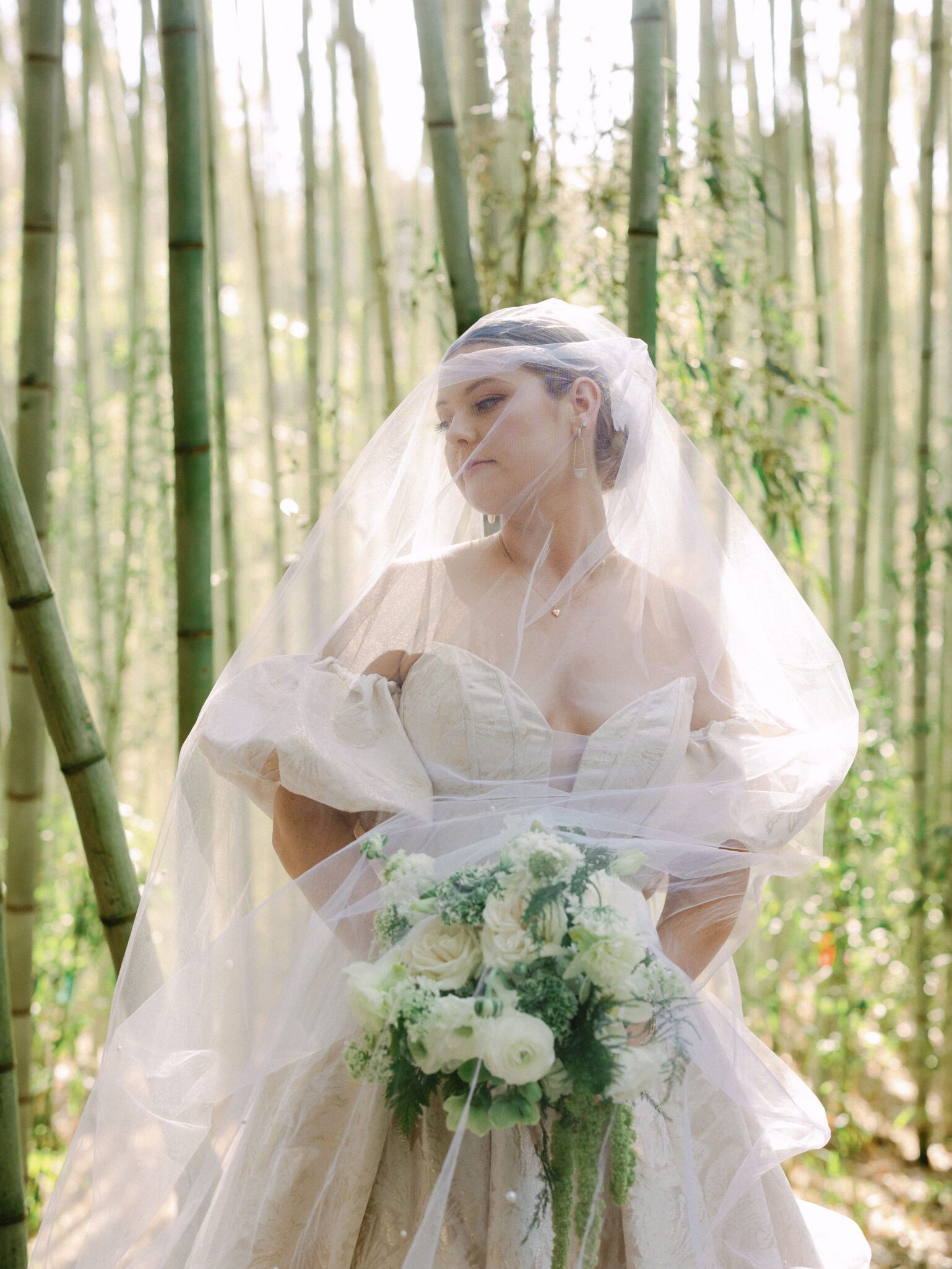 A bride in an elegant off-shoulder gown and veil stands in a sunlit bamboo forest at Camelot Meadows, Charlotte NC, holding a bouquet of white and green flowers. Dappled sunlight filters through the tall bamboo, evoking the charm of 35mm film.