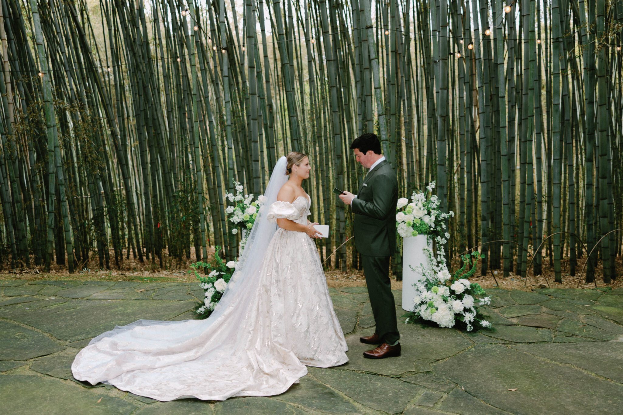 A bride in a white gown and veil stands facing a groom in a dark suit as he reads vows, surrounded by lush bamboo and white floral arrangements on a stone pathway.