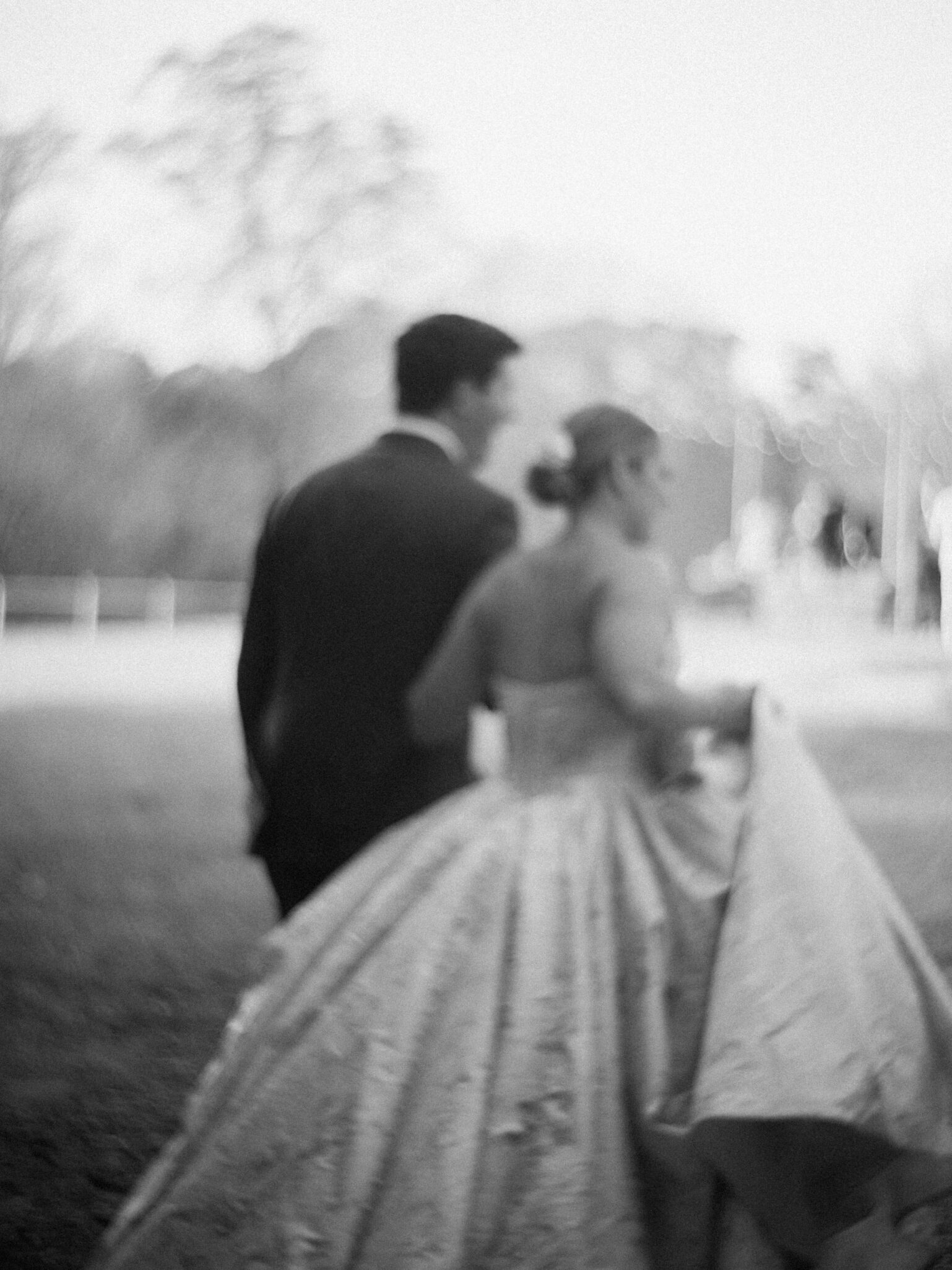 A black and white, blurred photo of a couple walking outdoors at Camelot Meadows. Shot on 35mm film in Charlotte, NC, the woman wears a large gown and the man is in a suit; their faces and the soft background remain indistinct.