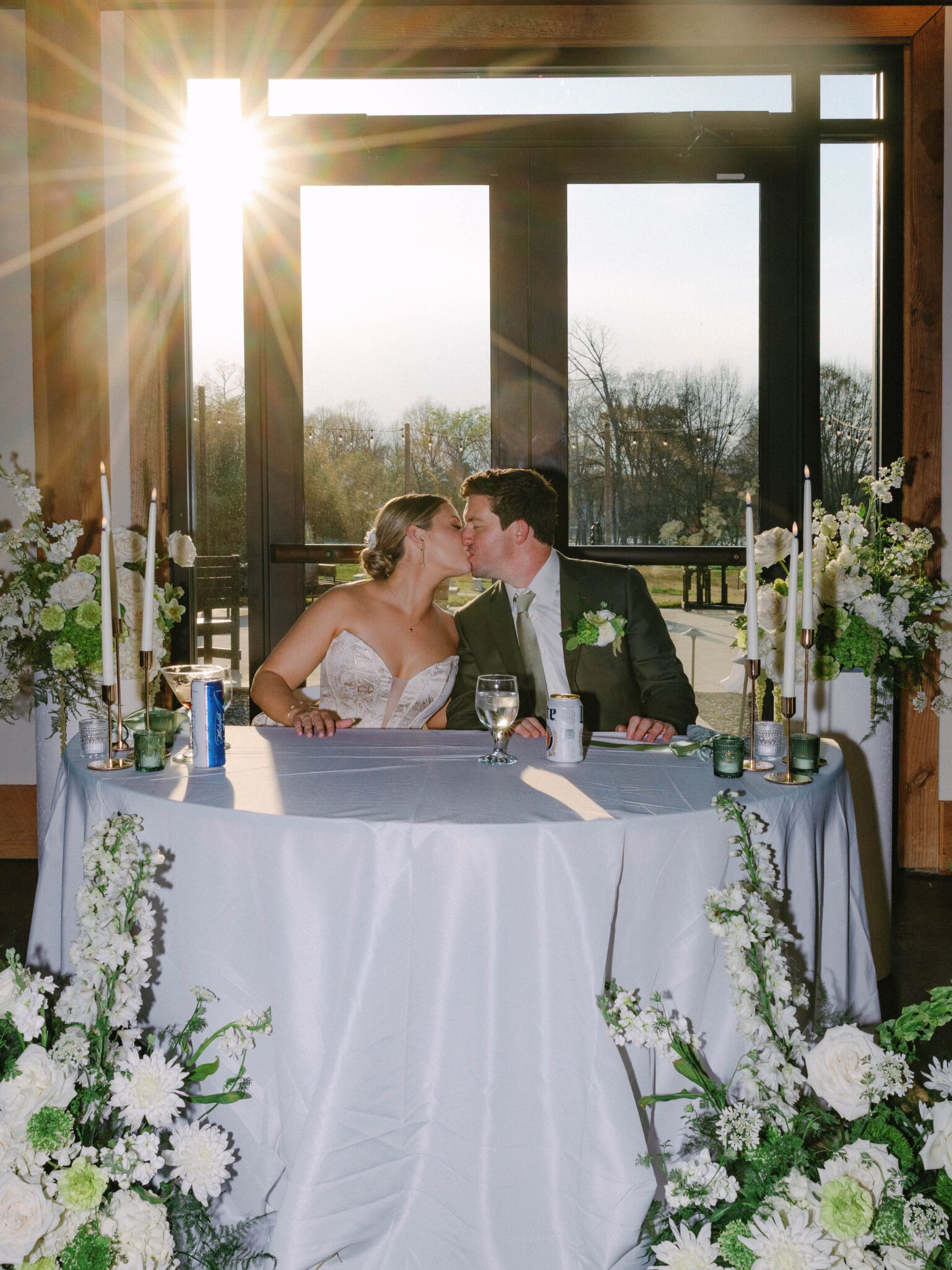 A newlywed couple sits at a decorated table, kissing, with sunlight streaming through large windows behind them at Camelot Meadows. Captured on 35mm film in Charlotte NC, white flowers and candles create a truly romantic atmosphere.
