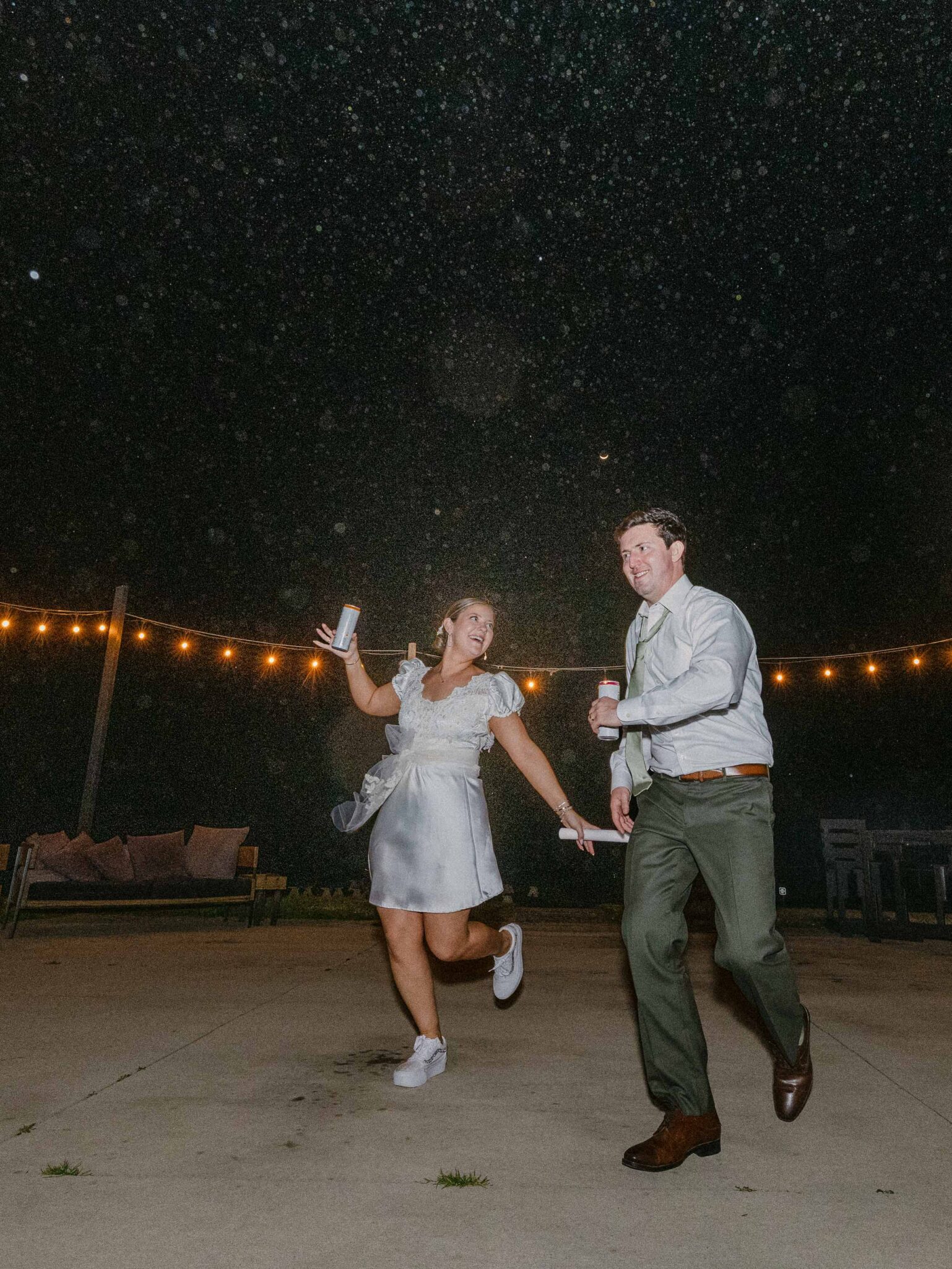 A woman in a short white dress and sneakers and a man in a white shirt and dark pants joyfully dance outdoors at night, holding drinks, with string lights and sprinkled rain illuminating the background.