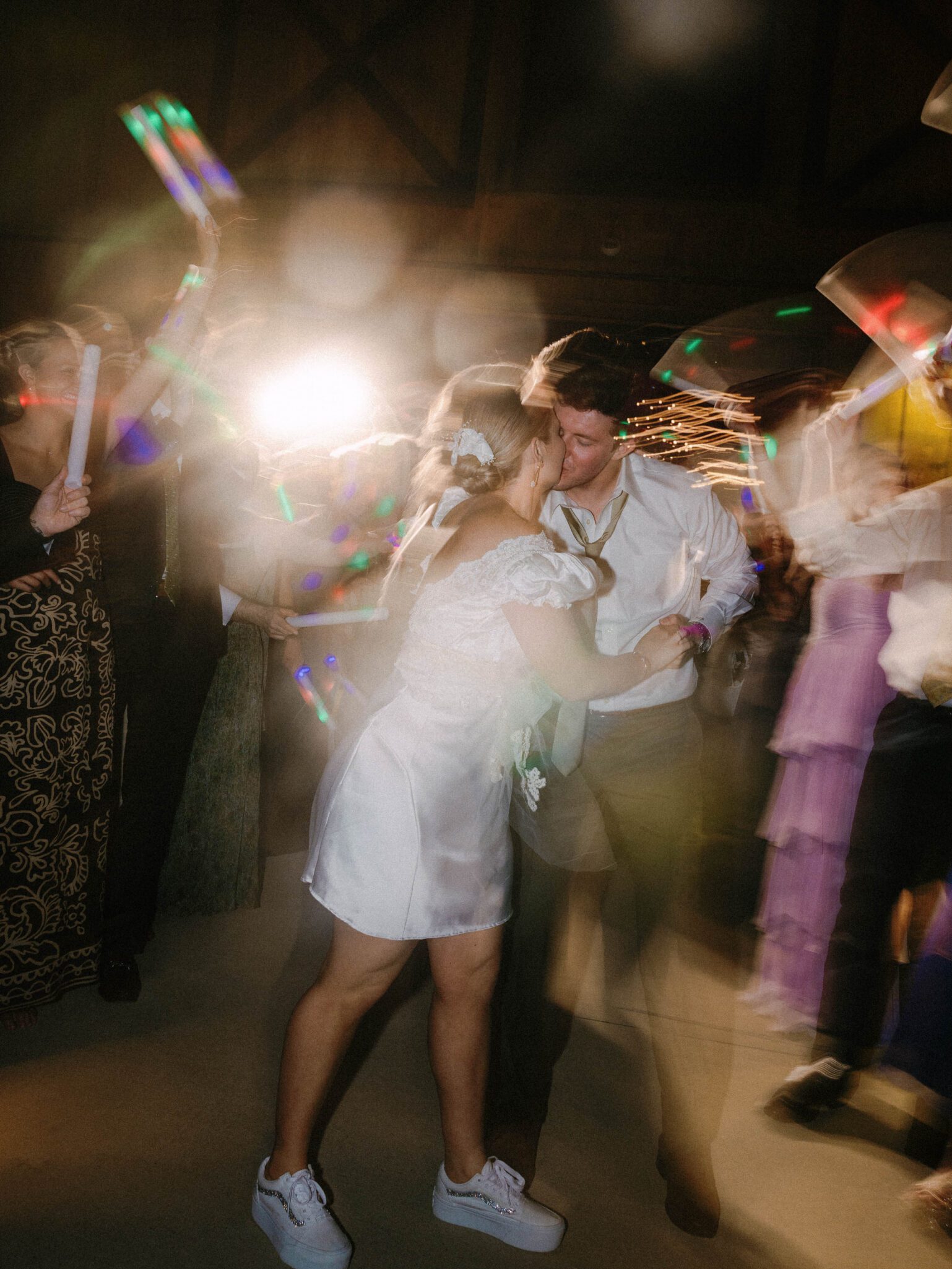 A couple dances closely at a lively Camelot Meadows party in Charlotte, NC, surrounded by blurred, moving guests and colorful lights. Captured on 35mm film, the joyful scene features her white dress and sneakers and his shirt and tie.
