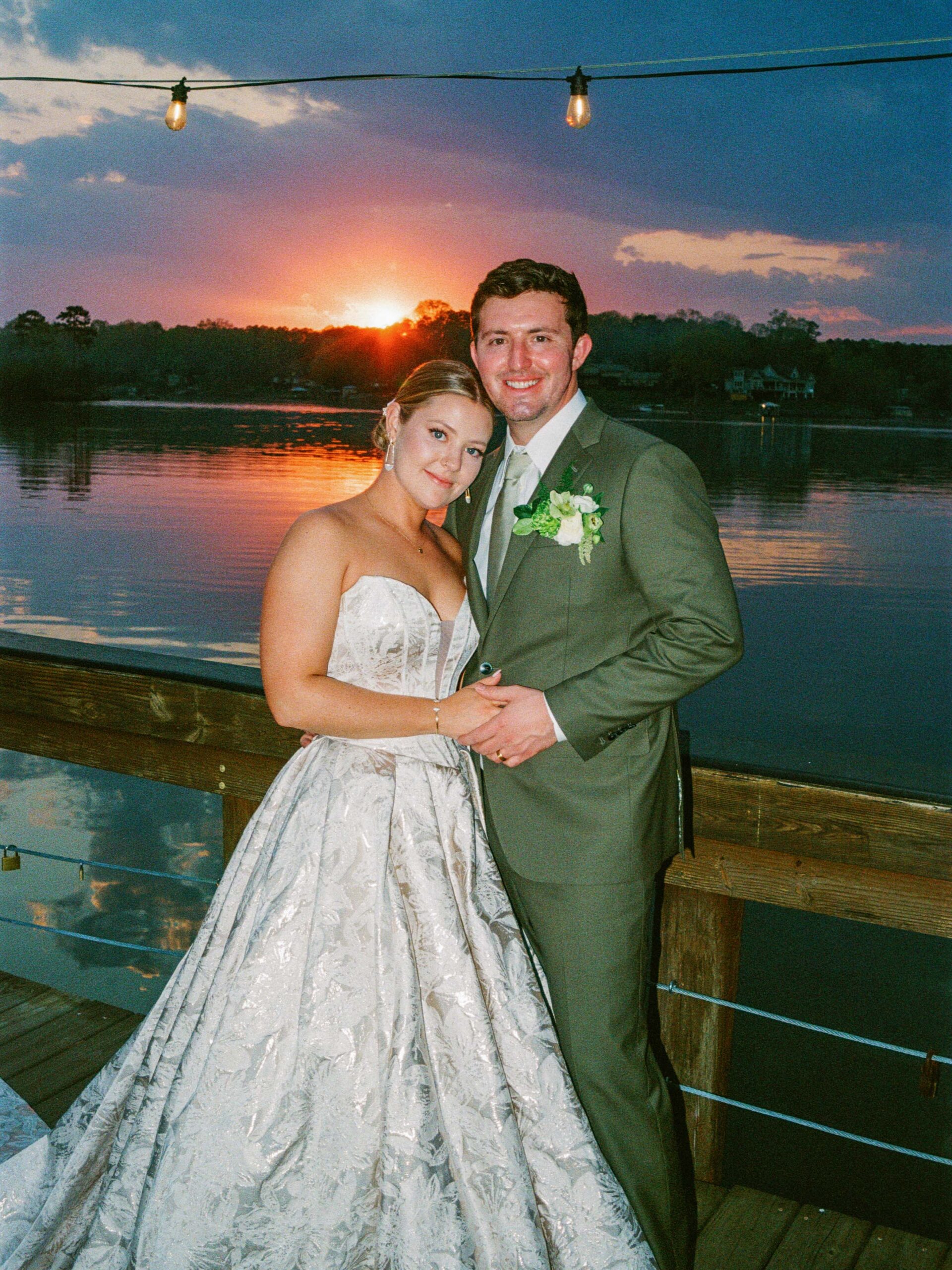 A bride in a strapless white gown and a groom in a green suit stand together on a wooden deck by a lake at sunset, smiling and embracing under string lights.