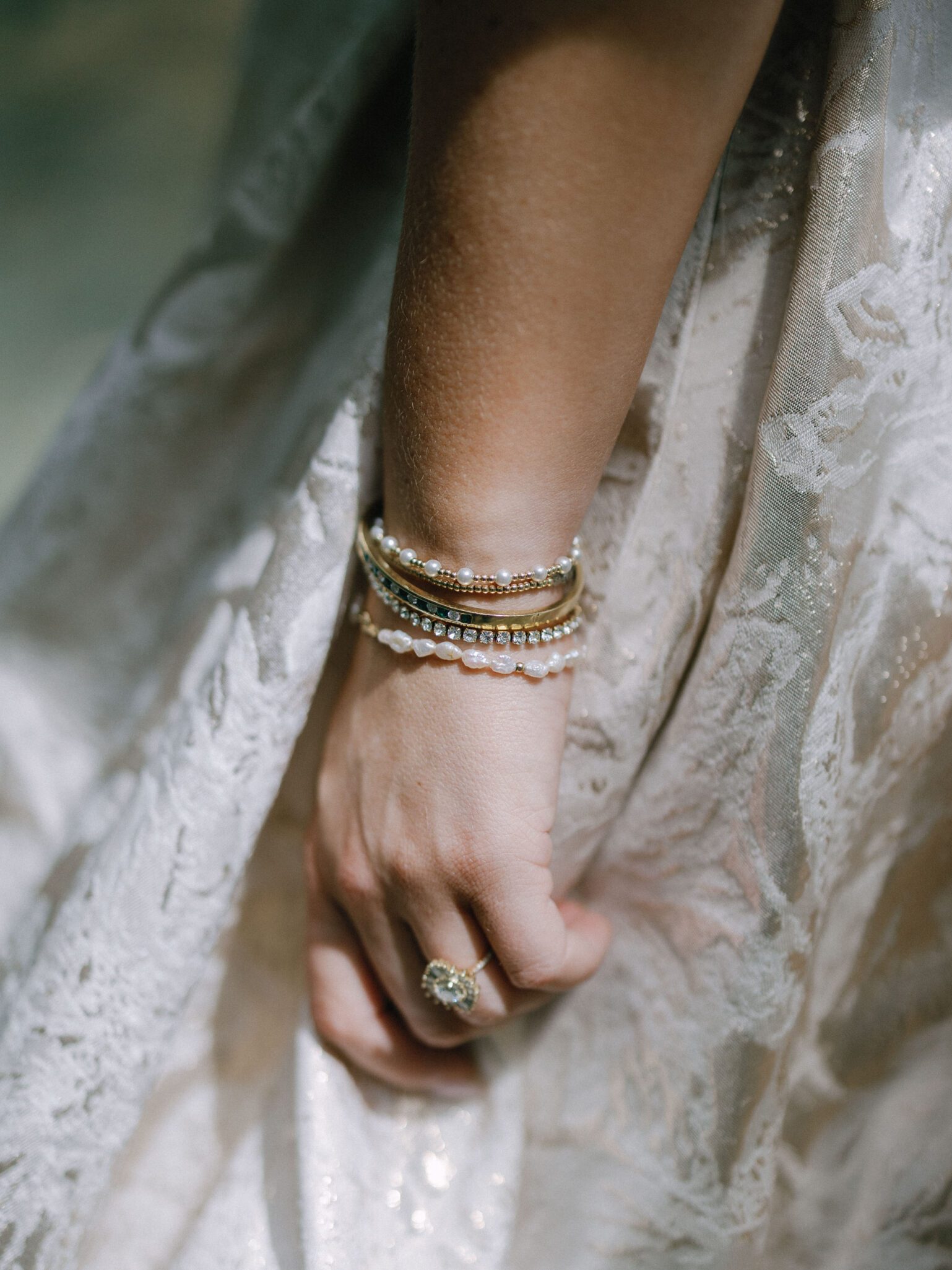 A close-up of a persons hand wearing gold and pearl bracelets and a vintage-style ring, resting against a light, lacy fabric dress.