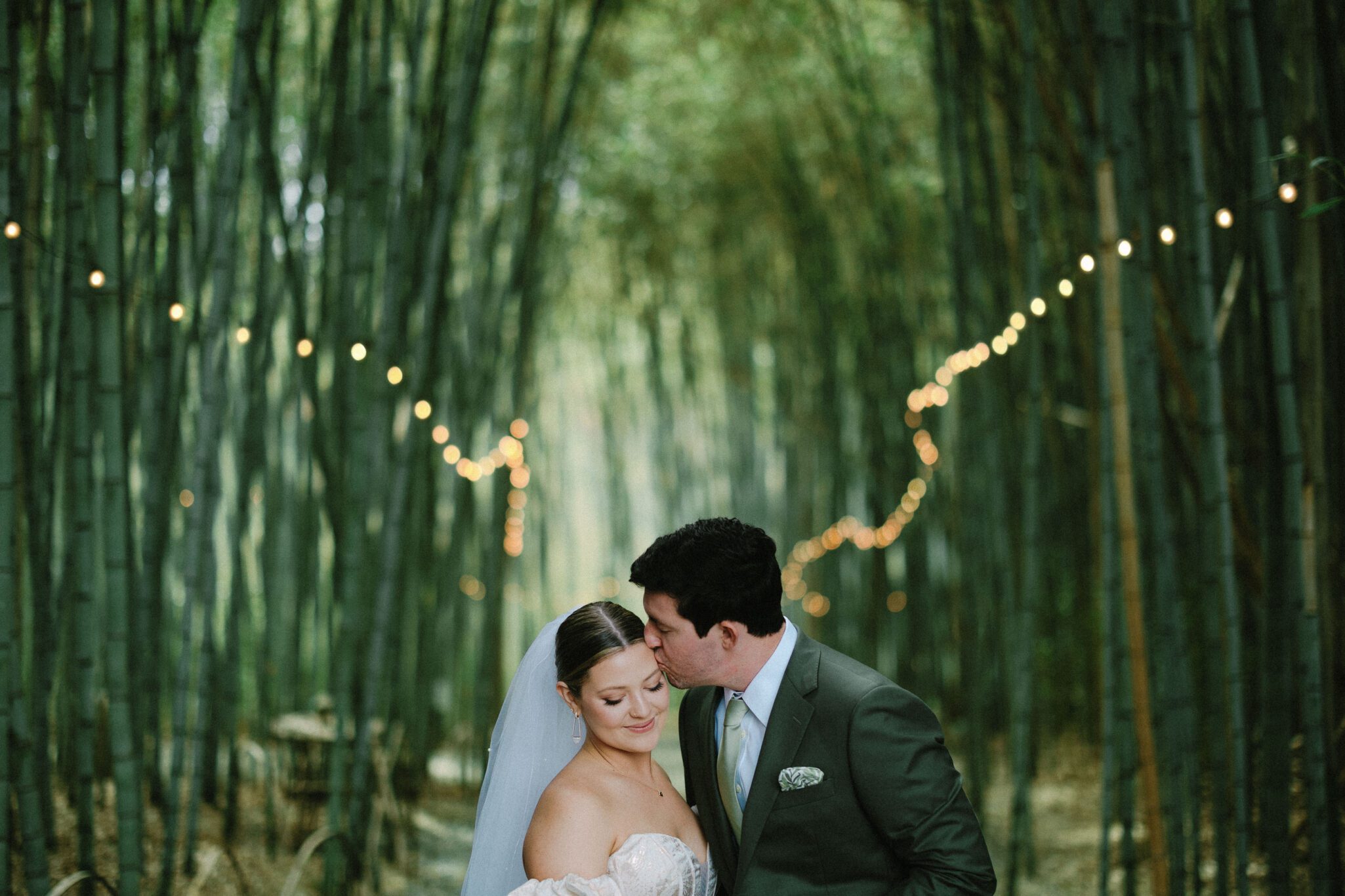 A groom in a dark suit kisses his brides forehead as she smiles with her eyes closed, standing together in a bamboo forest with string lights overhead.