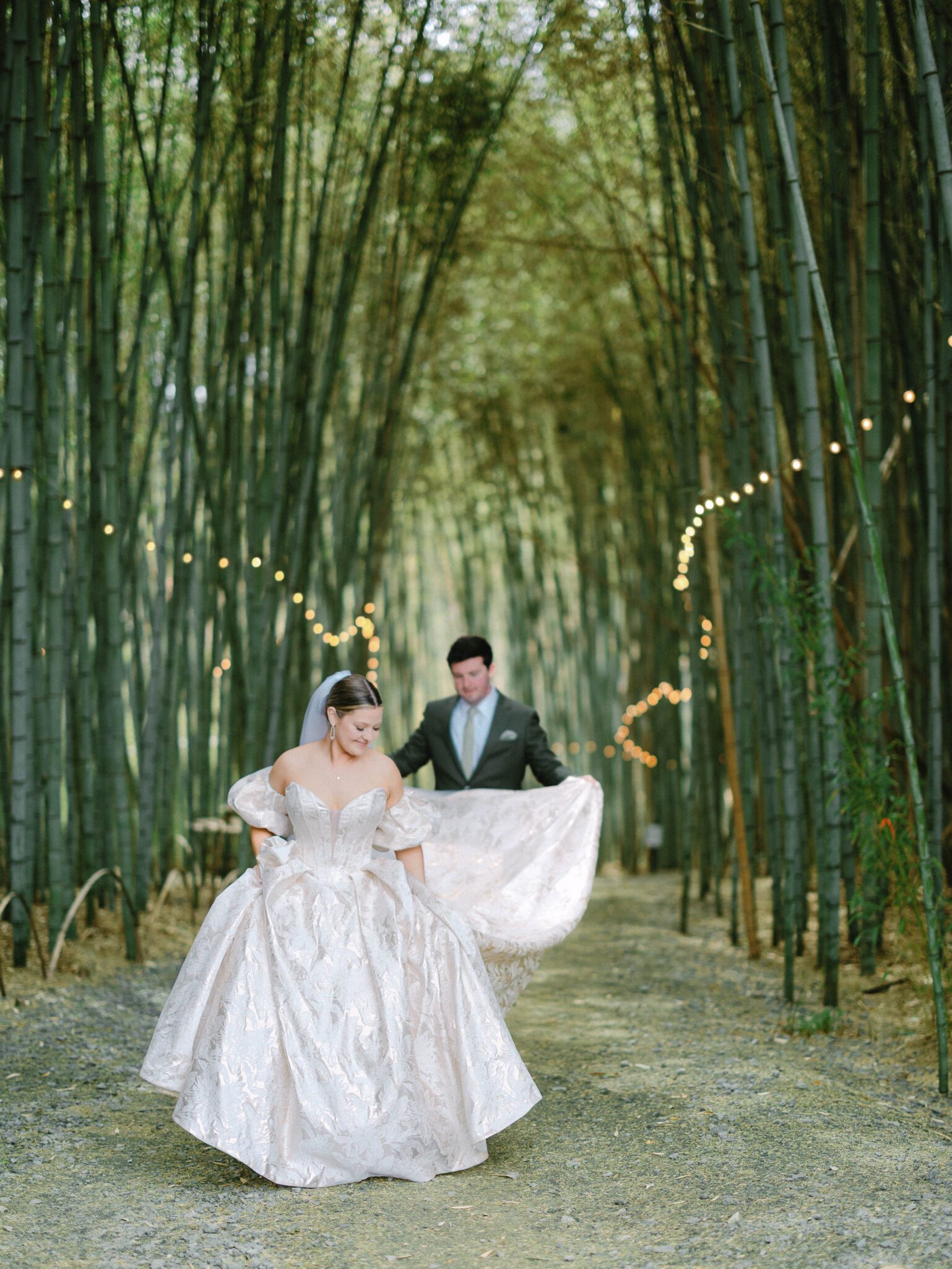A bride in a shimmering gown smiles as she walks through a bamboo forest, while the groom holds her train. String lights hang overhead, creating a magical, romantic atmosphere.