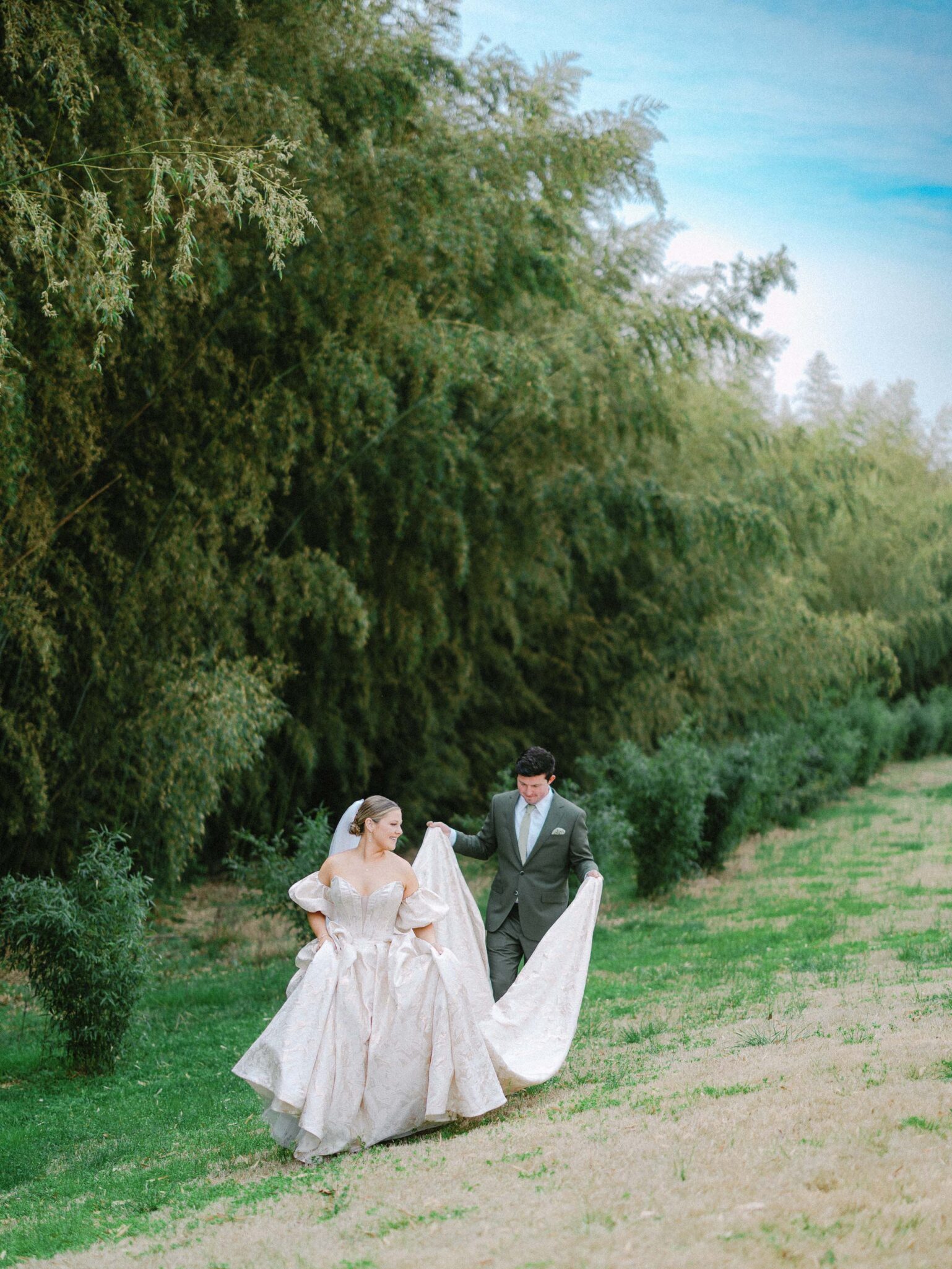 A bride in a white gown and veil walks on grass while a groom in a gray suit holds her dress train. They are outdoors with tall green trees and a blue sky in the background.