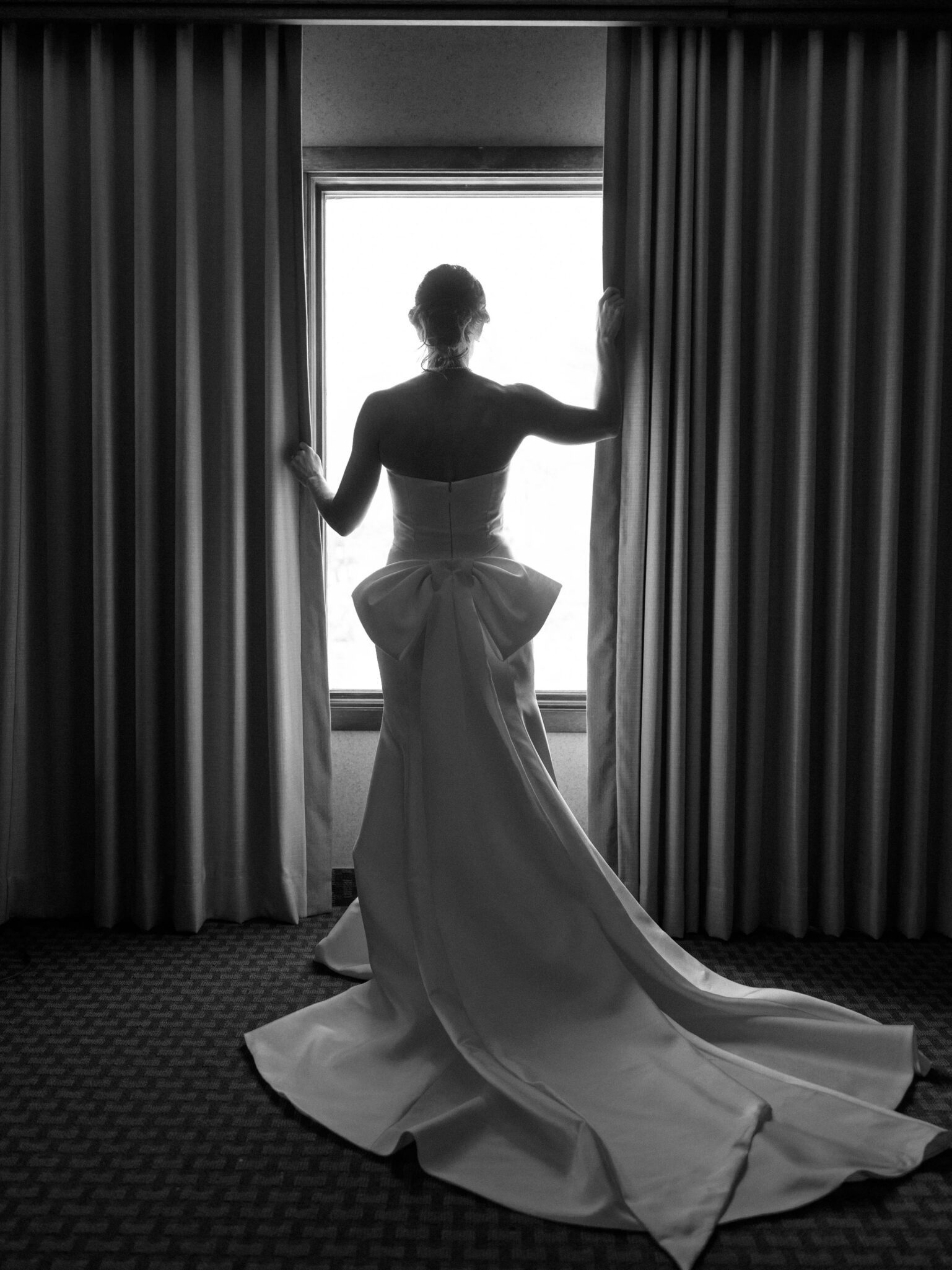 A woman in a long, elegant wedding dress with a large bow stands facing a window, gently holding open the curtains. Daylight streams in, silhouetting her figure in a softly lit room.