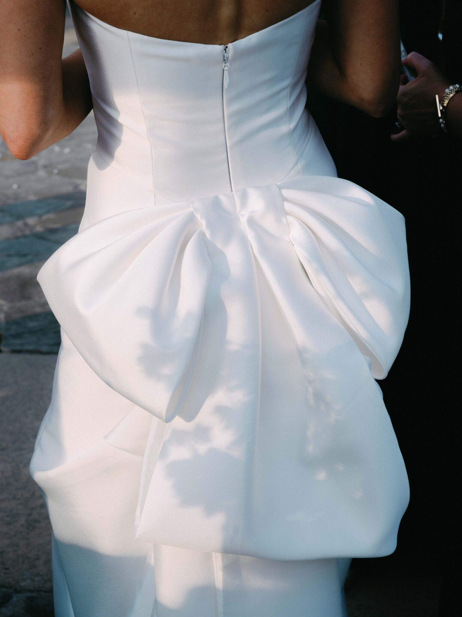 A close-up of a person in a white strapless dress featuring a large, elegant bow on the back, with soft sunlight creating subtle shadows on the fabric.