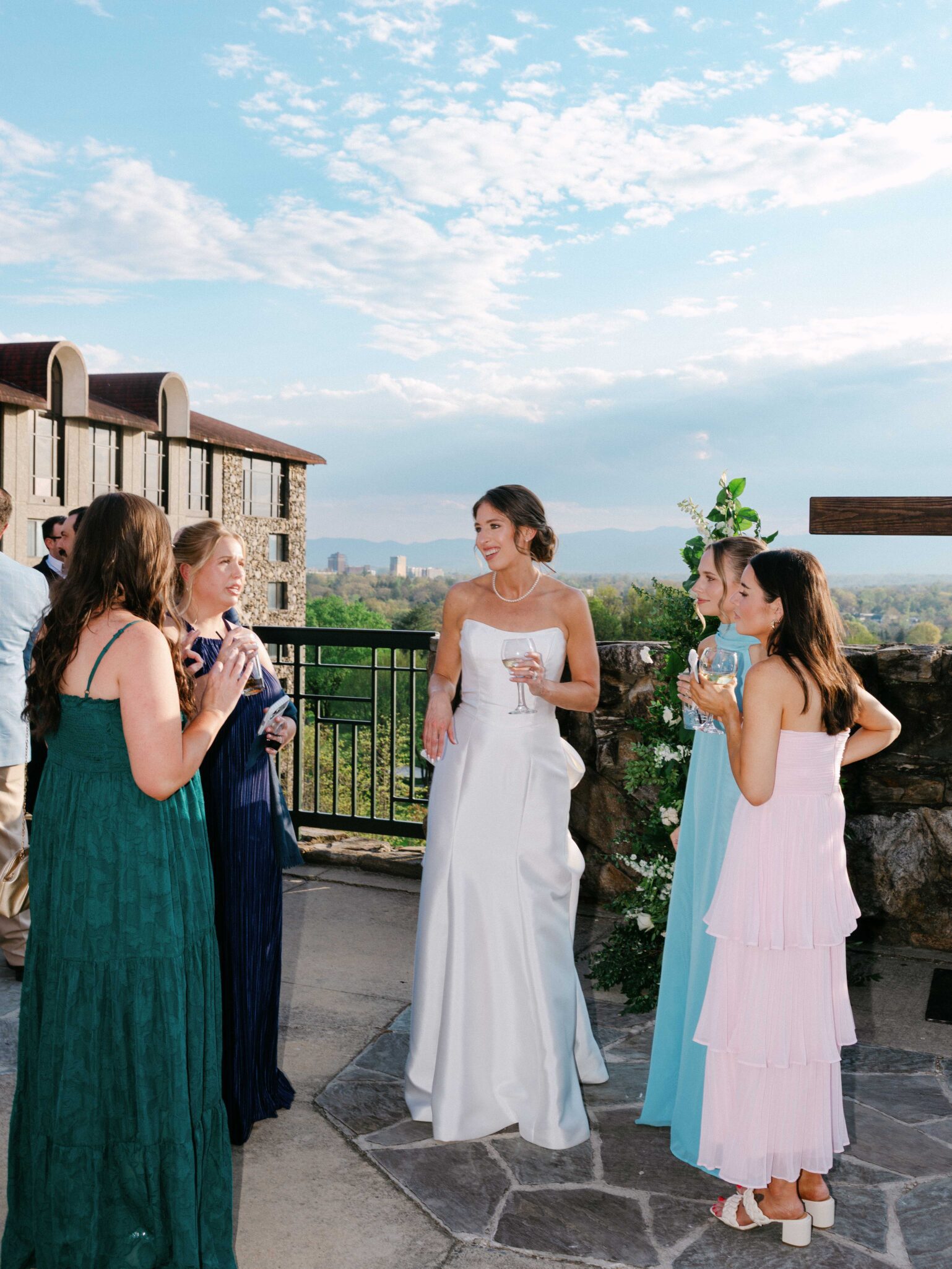 A bride in a white dress stands outside with four women in colorful dresses, all holding drinks and talking, as scenic mountains and a blue sky set the backdrop—captured beautifully by The Omni Grove Park Inn wedding photographer.