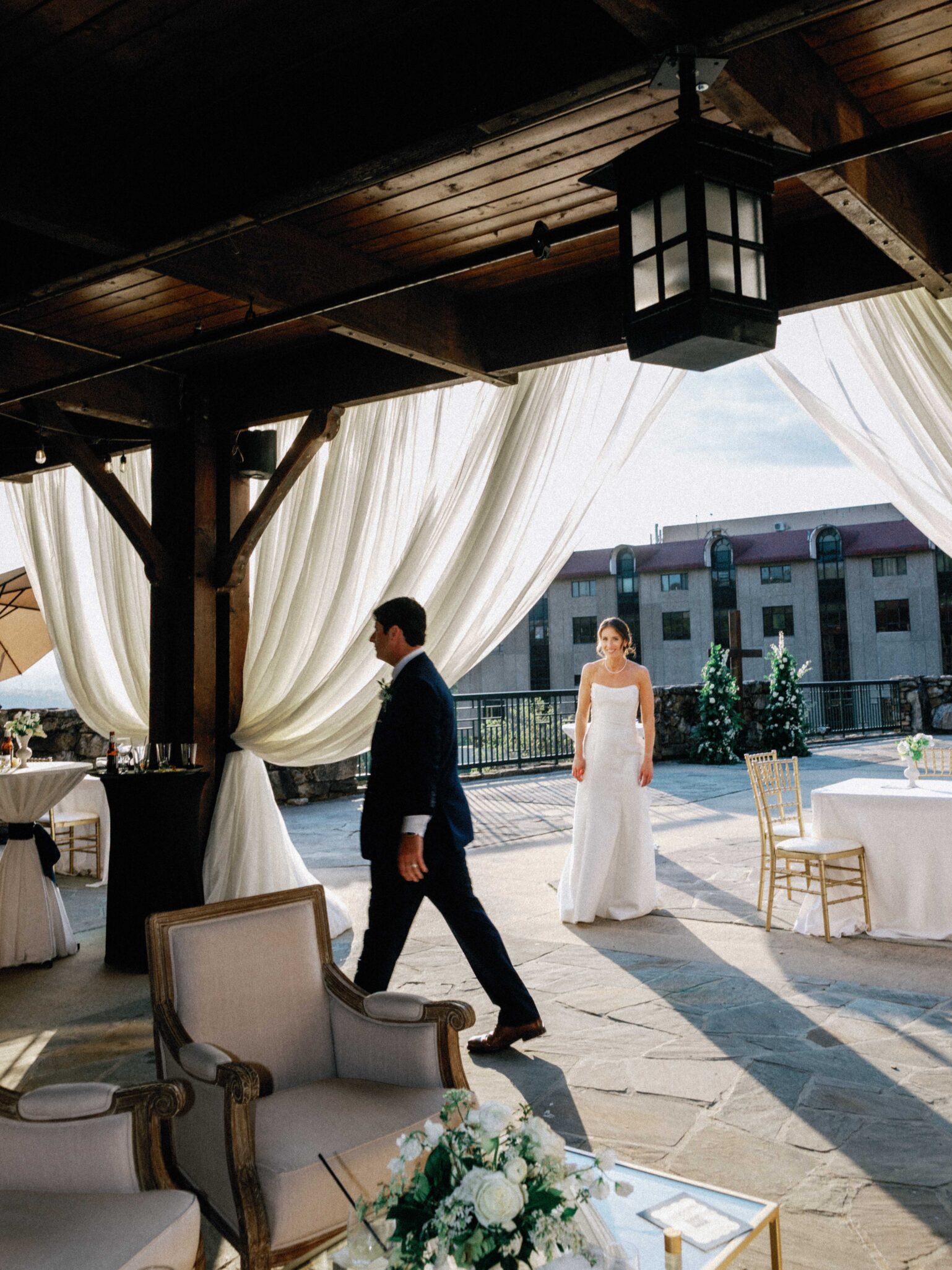 A bride in a white gown walks toward a groom in a navy suit under a wooden pavilion with white drapes, elegant chairs, and tables, with a building and greenery visible outside.