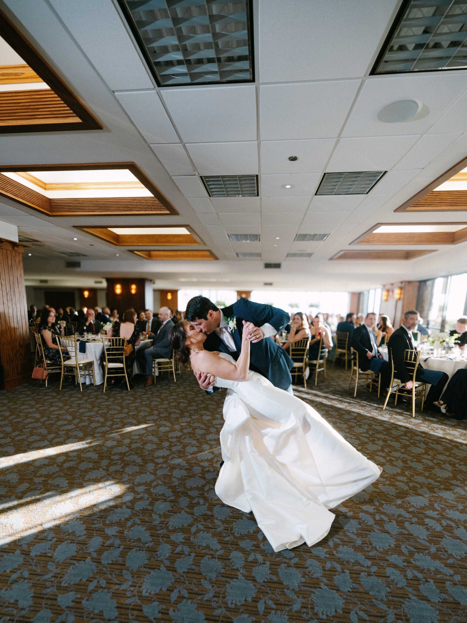 A bride and groom share a romantic dip during their first dance at a wedding reception, surrounded by seated guests in a bright, elegant banquet hall.