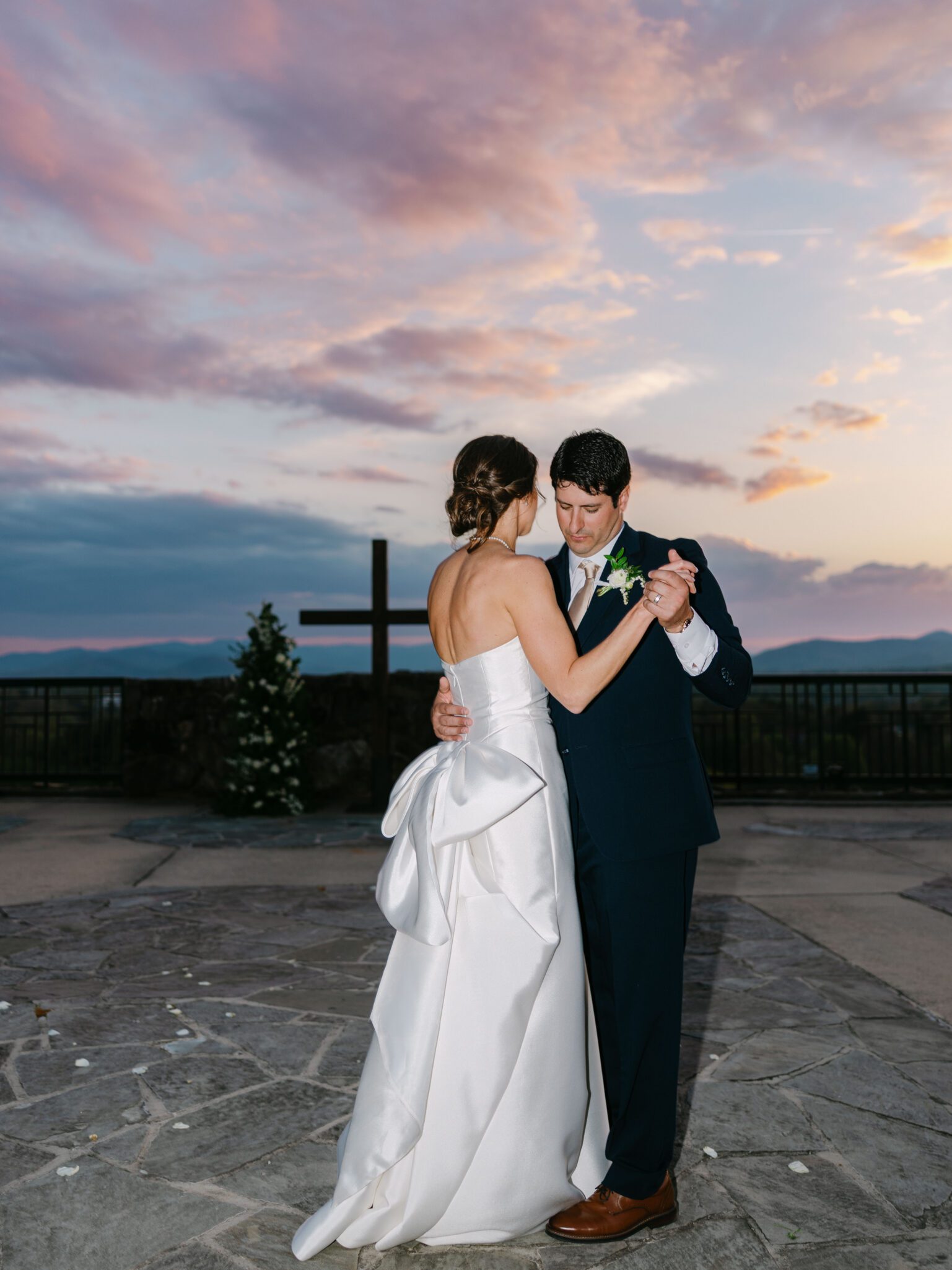 A bride in a white gown and a groom in a dark suit share a dance outdoors at sunset, with a cross and mountains in the background under a colorful sky.