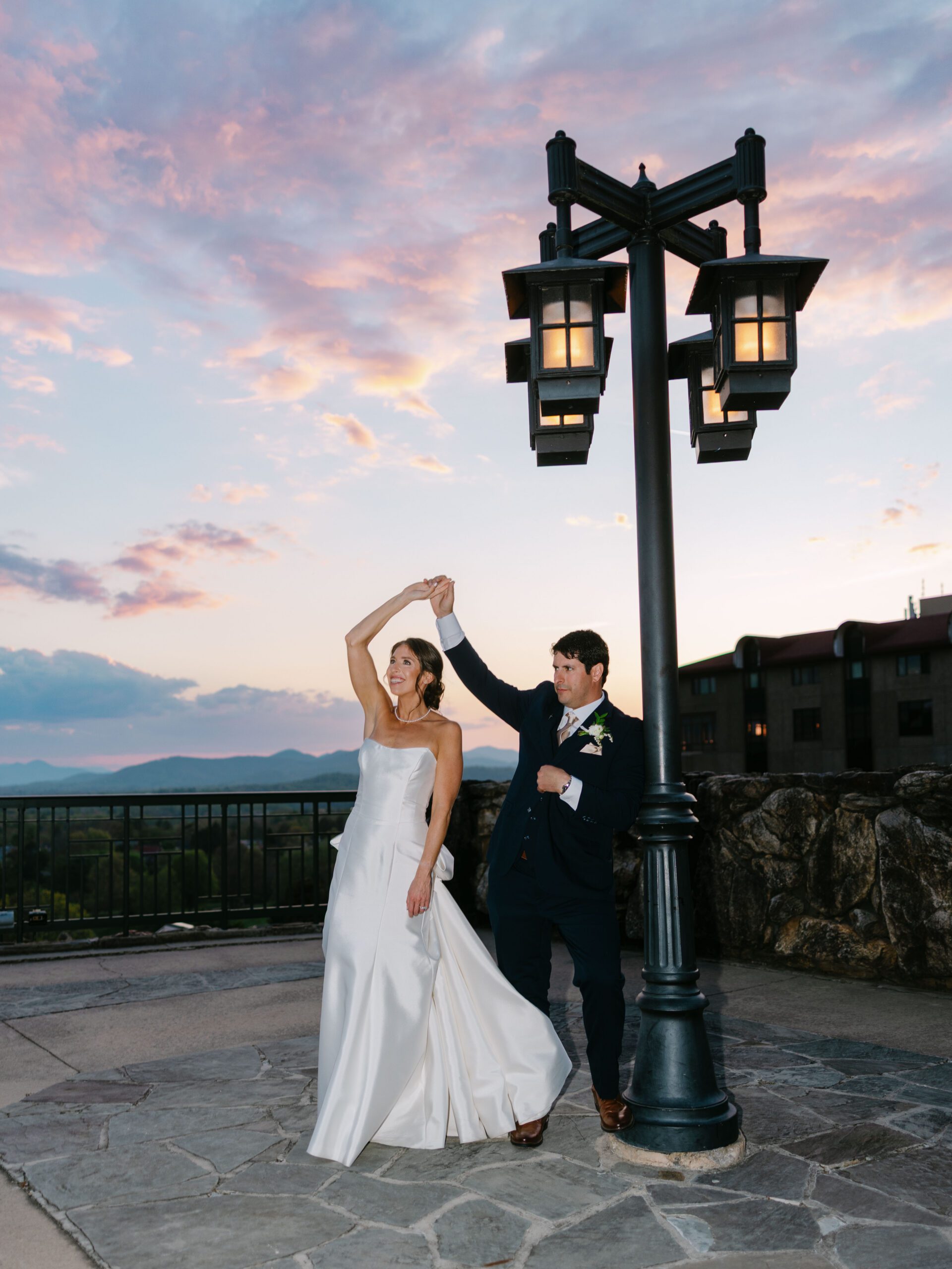 A bride and groom dance outdoors at sunset under a streetlamp. Captured by The Omni Grove Park Inn wedding photographer, the bride in a white dress smiles as she’s twirled by her groom beneath colorful pink and purple clouds.