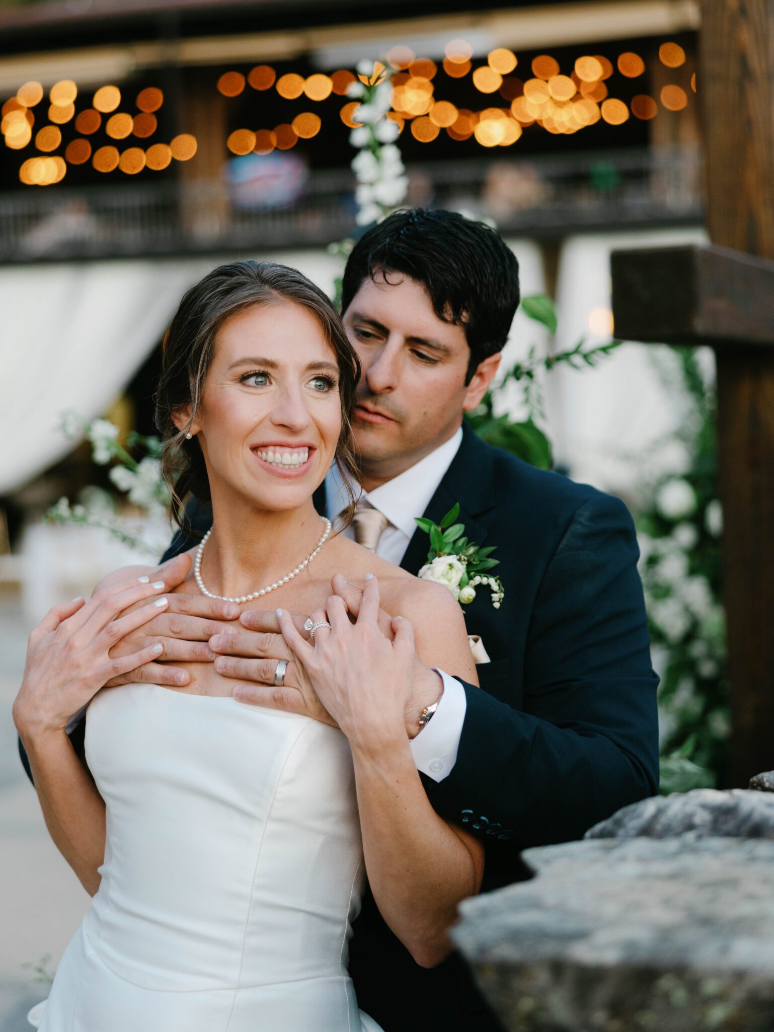A bride in a white dress and pearl necklace smiles while a groom in a dark suit stands behind her, embracing her. String lights and greenery decorate the background, creating a warm, romantic atmosphere.