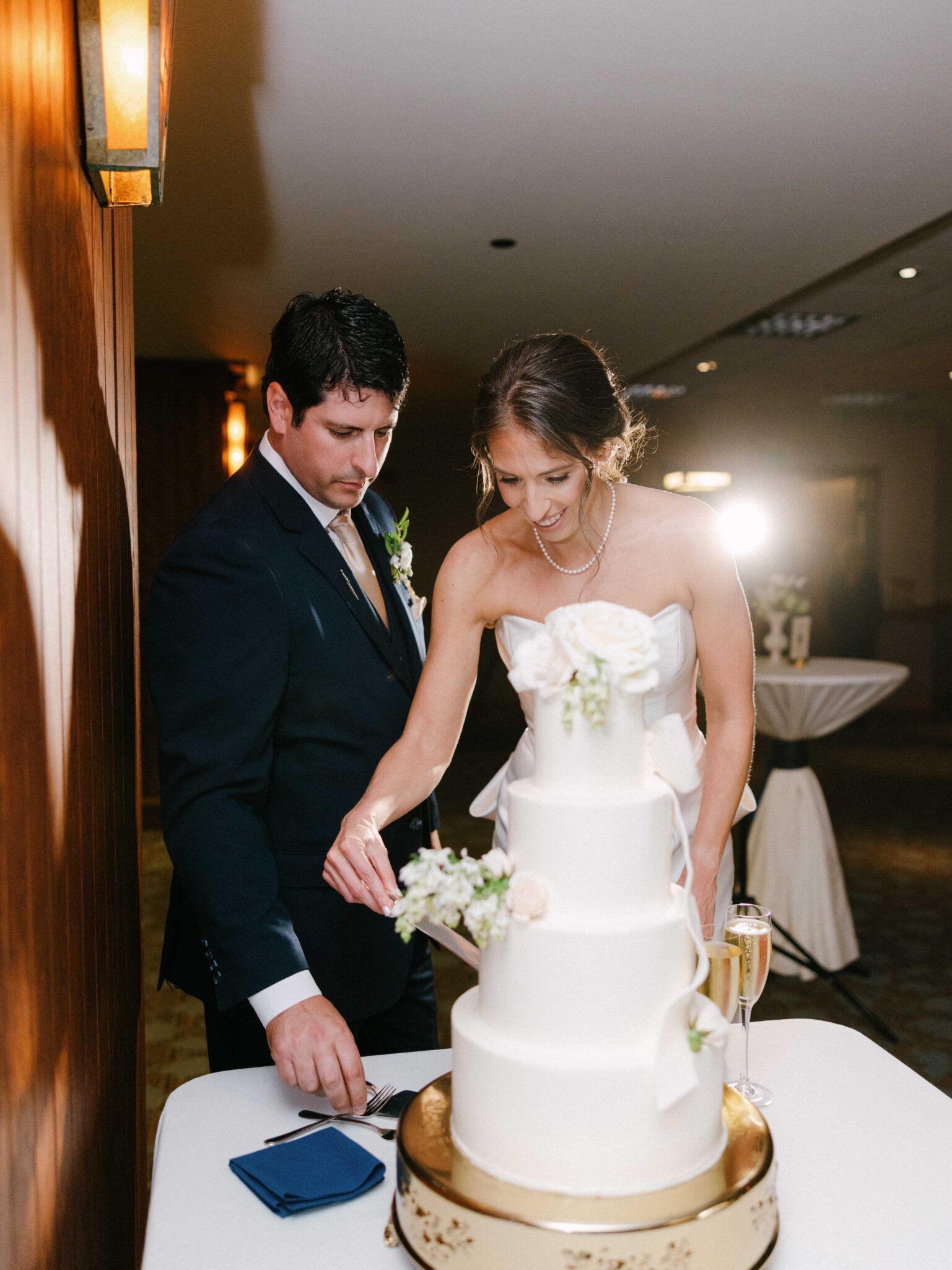 A bride and groom stand together, smiling as they cut a tall white wedding cake decorated with flowers. The bride wears a white dress and pearls; the groom is in a dark suit. A champagne glass sits on the table.