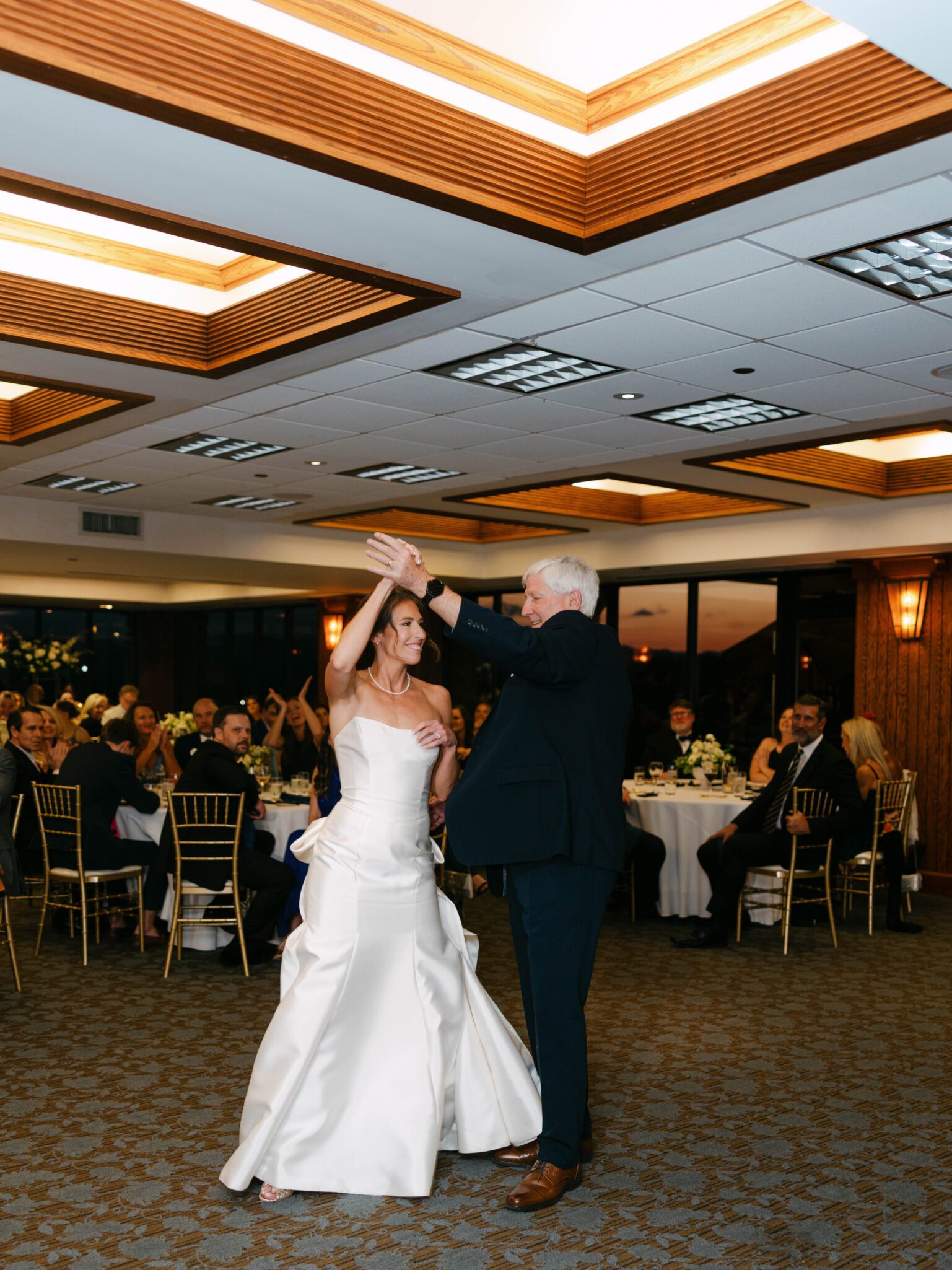 A bride in a white wedding gown dances with an older man in a suit on a ballroom floor, with guests seated at round tables watching and smiling in the background.