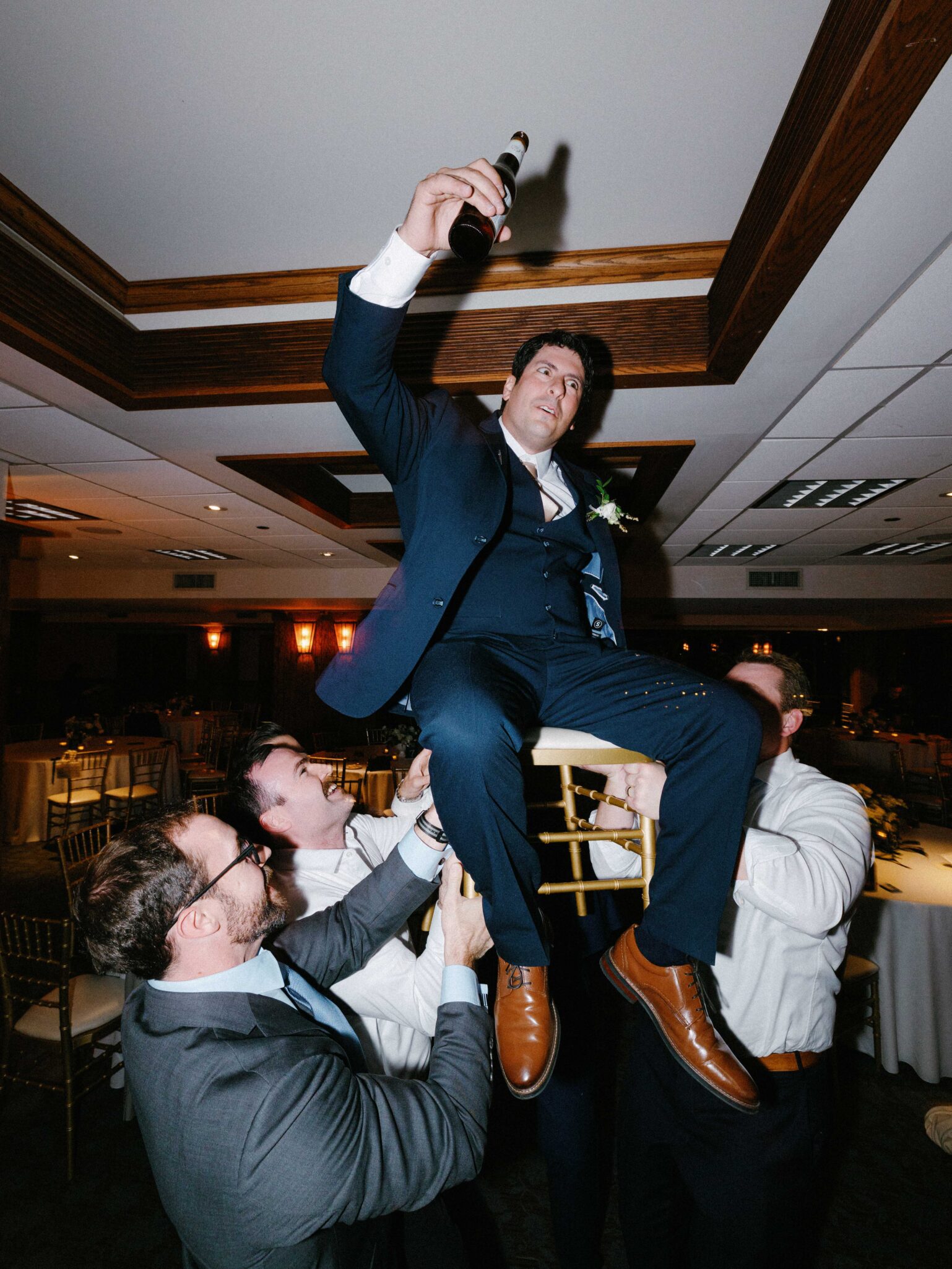 A man in a suit holds up a beer bottle while sitting on a chair being lifted by three other men at an indoor celebration or party.