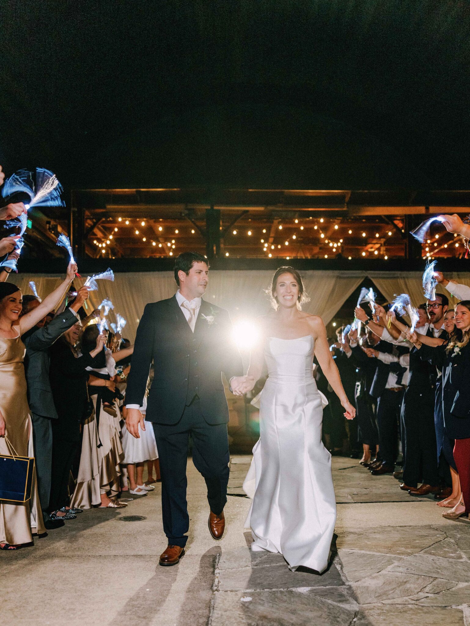 A bride and groom walk hand in hand at night, smiling as guests cheer and wave streamers. String lights illuminate the background, creating a festive atmosphere.