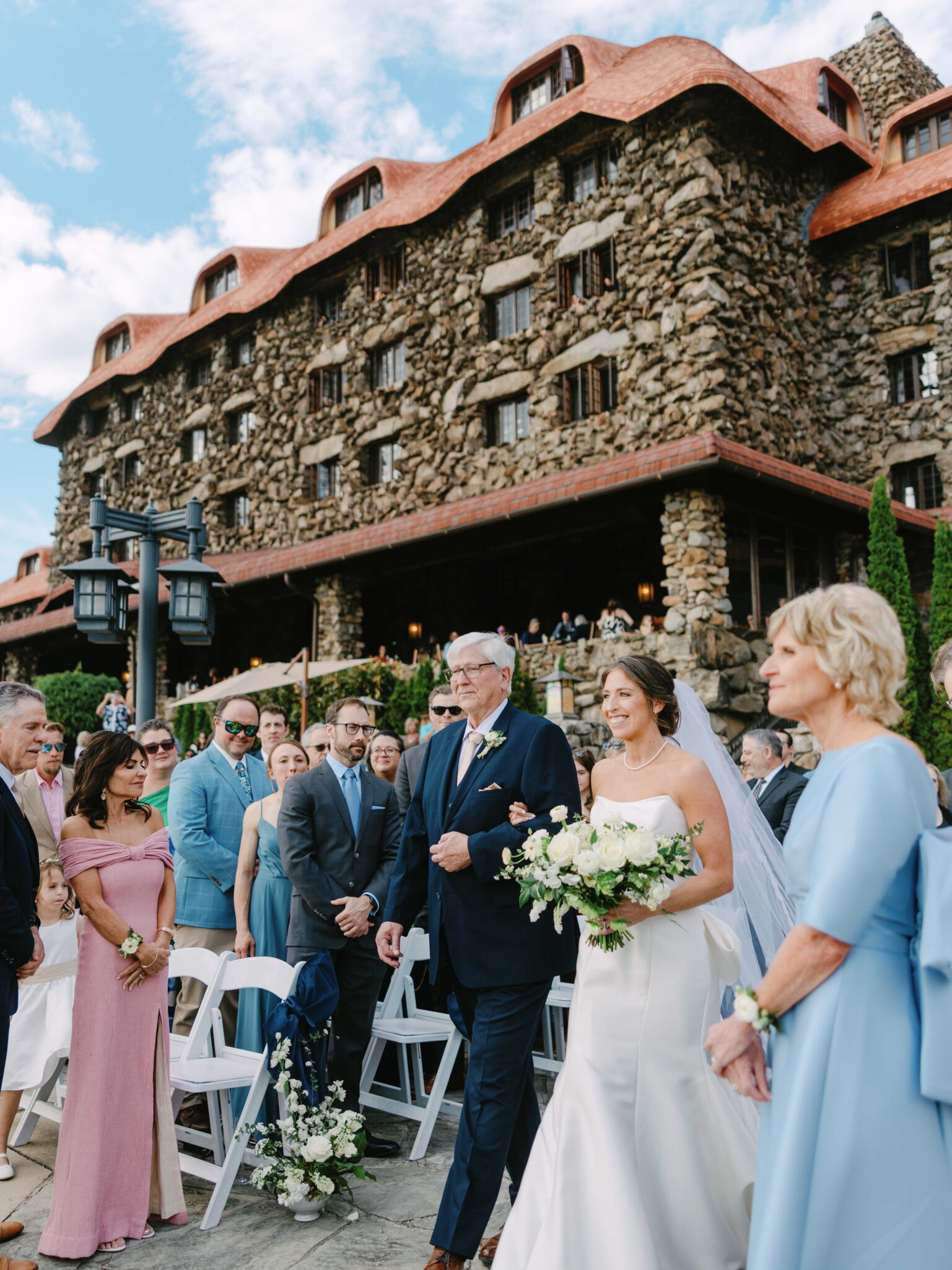 A bride in a white dress and veil walks down the aisle with an older man, surrounded by guests in formal attire, in front of a large stone building with a red roof on a sunny day.