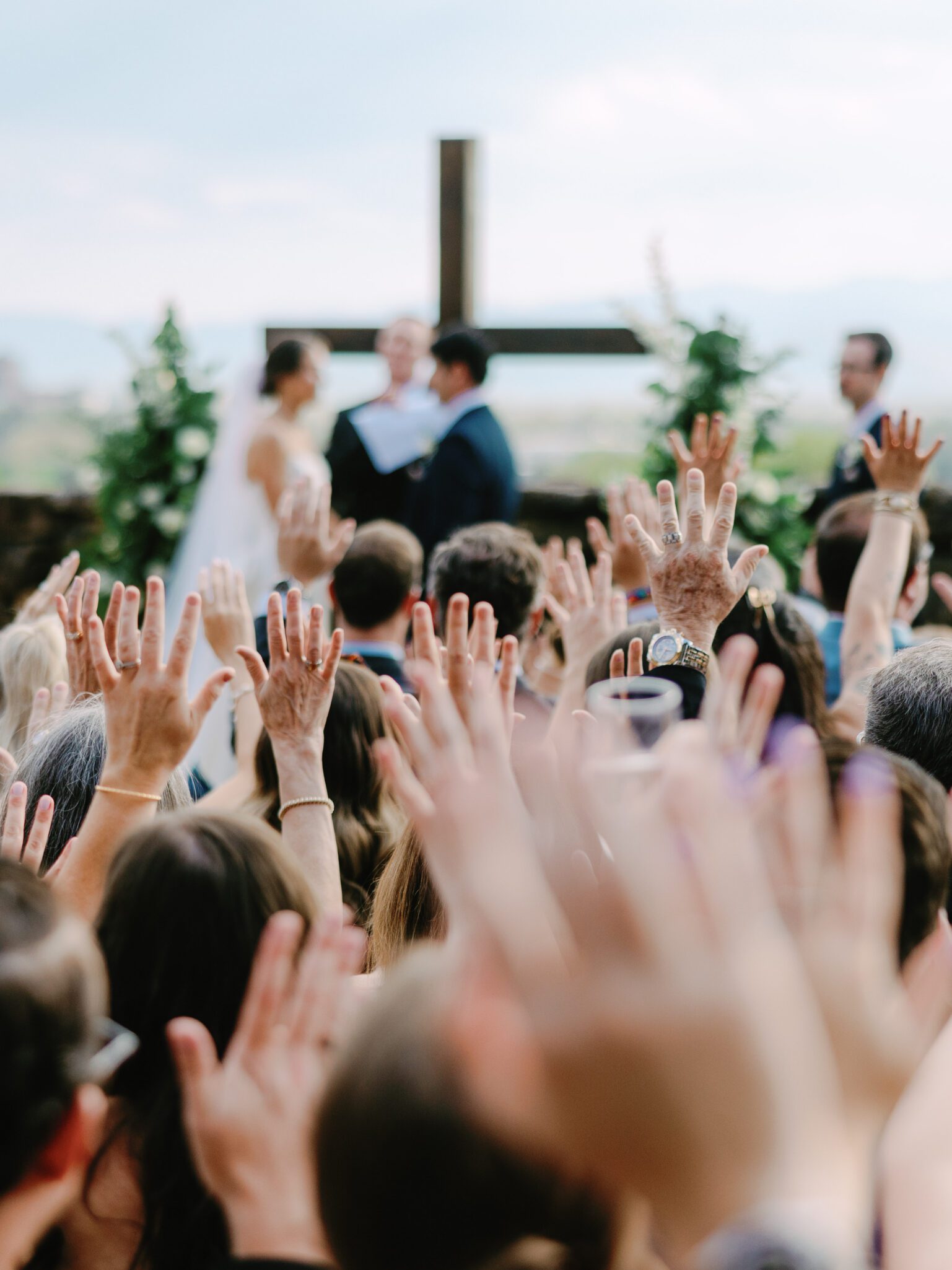 A crowd of people with raised hands facing a bride and groom standing before a large cross during an outdoor wedding ceremony.