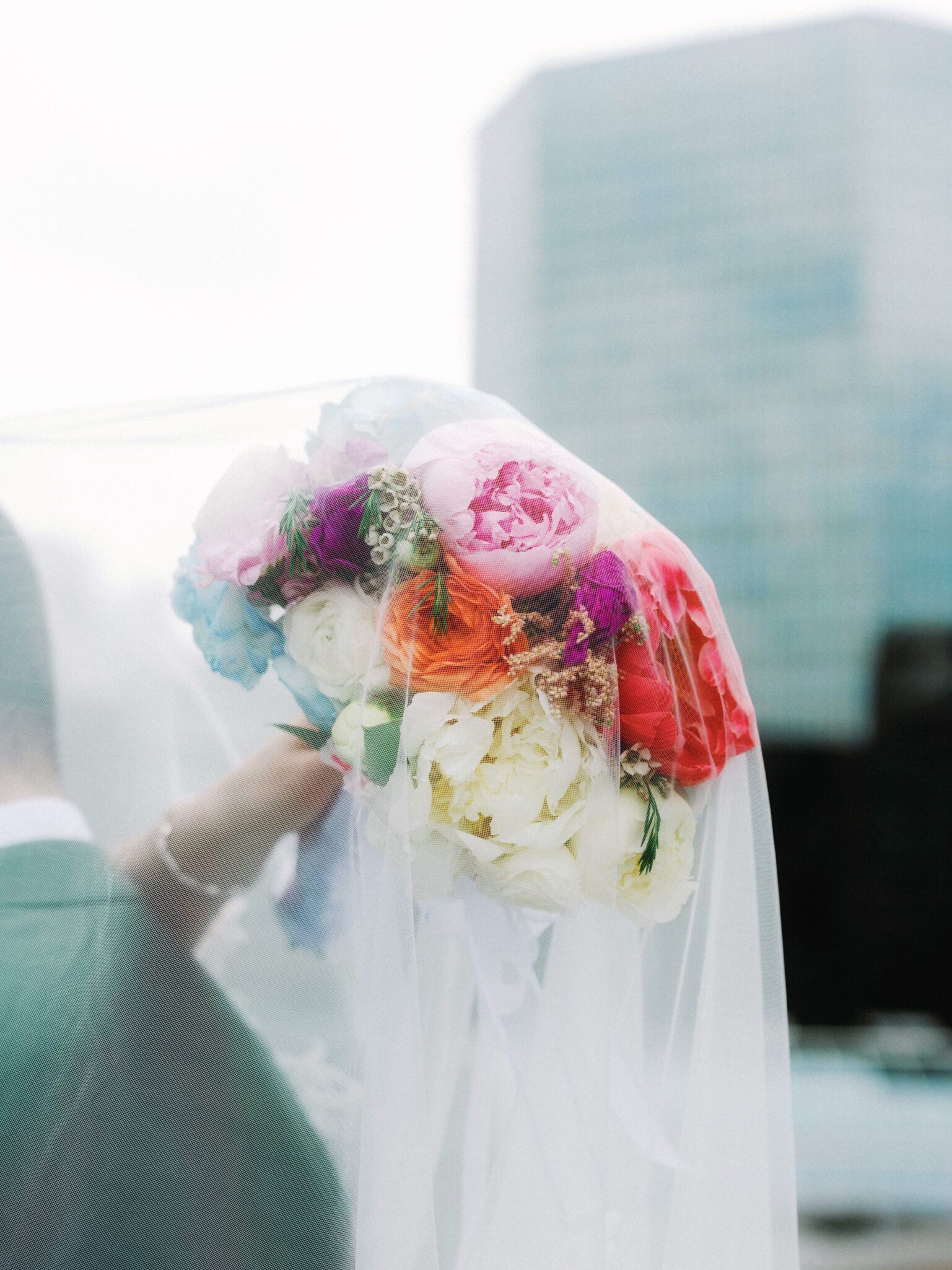 A person in a suit holds a colorful bouquet of flowers wrapped in white tulle, with tall, blurred glass buildings in the background.