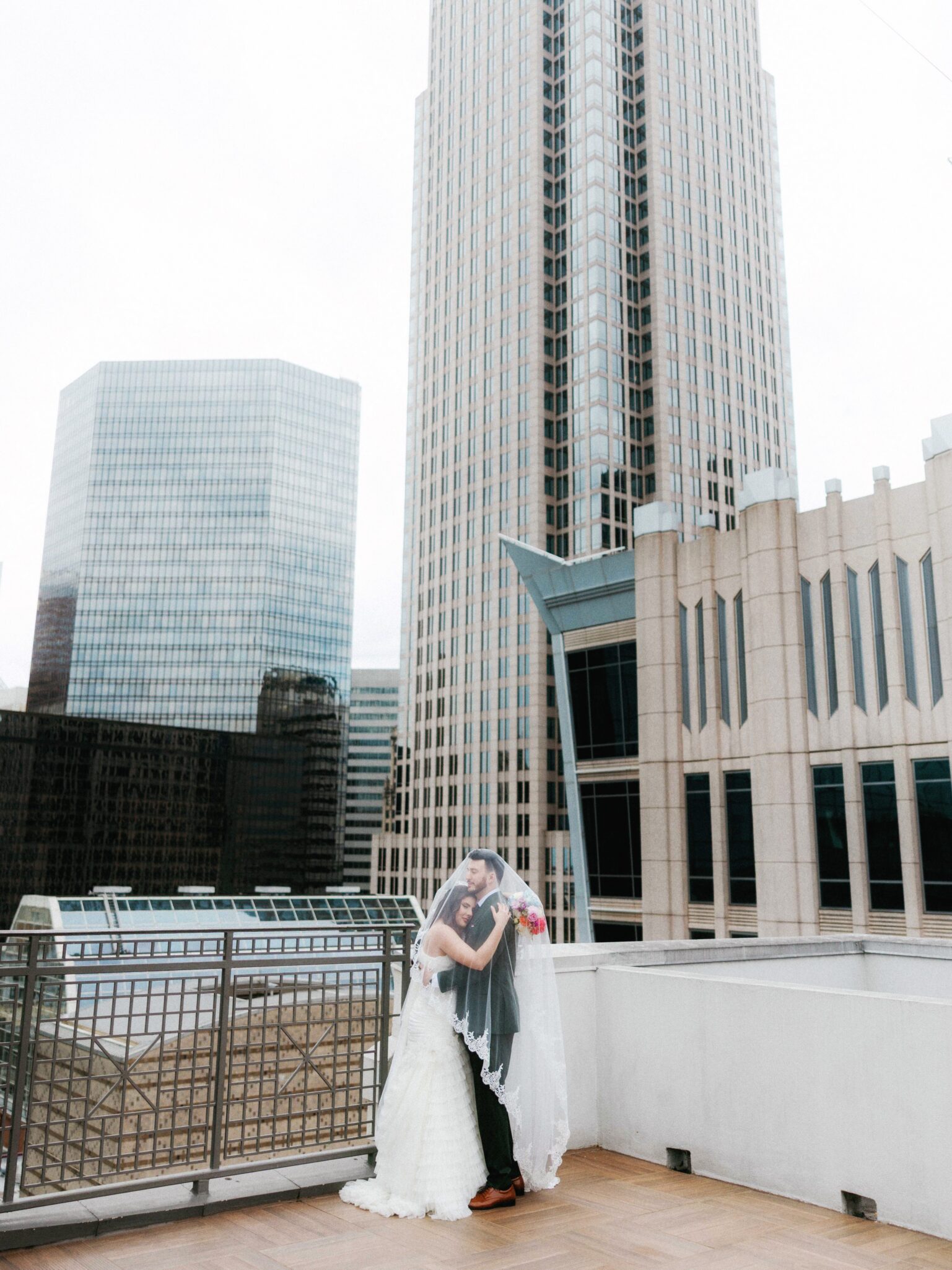 A bride and groom embrace on a rooftop at the Double Tree Hilton Charlotte City Center, covered by a bridal veil, with tall modern skyscrapers in the background. The scene is bright and urban, capturing a romantic moment amid a cityscape.