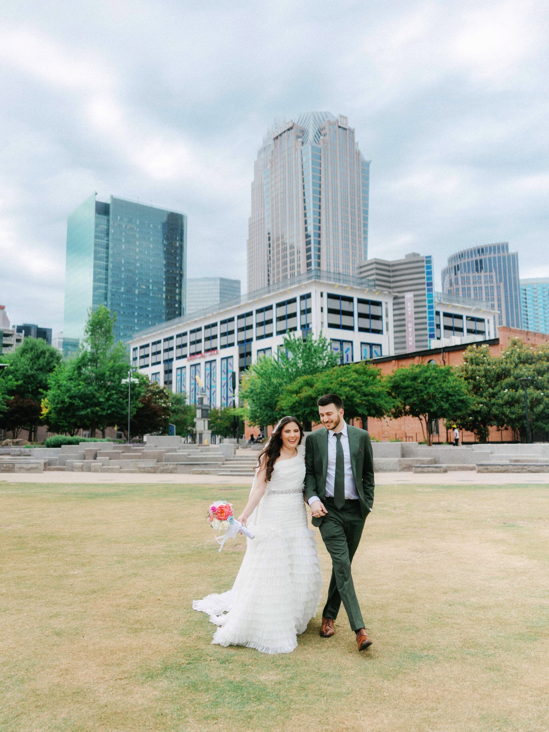 A bride in a white dress and a groom in a dark suit walk hand in hand on a grassy lawn, smiling, with tall modern buildings and the Double Tree Hilton Charlotte City Center Wedding venue under a cloudy sky in the background.