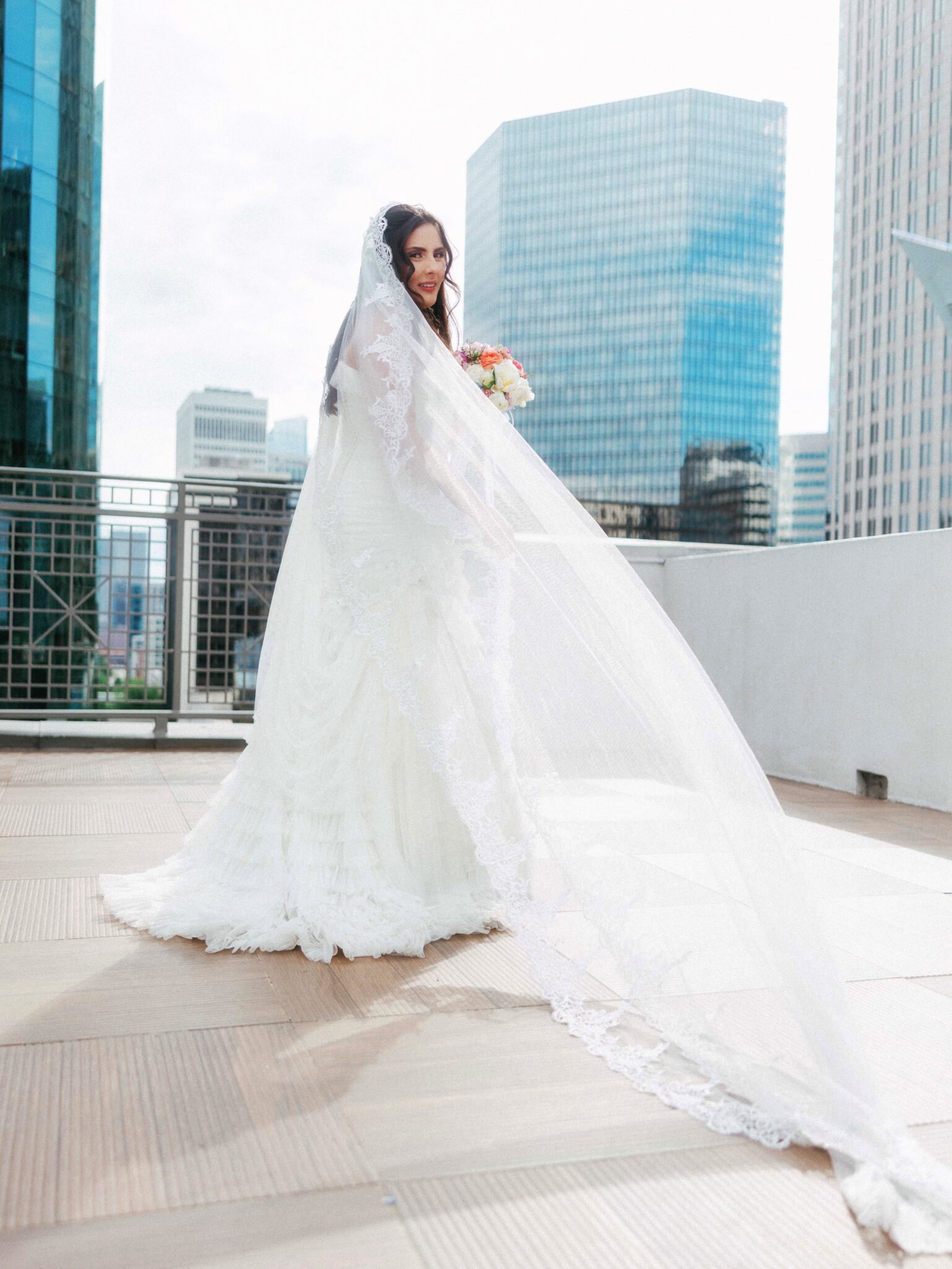 A bride in a long white wedding gown and lace veil stands on a rooftop at the DoubleTree Hilton Charlotte City Center, holding a bouquet of flowers, with modern city skyscrapers in the background.