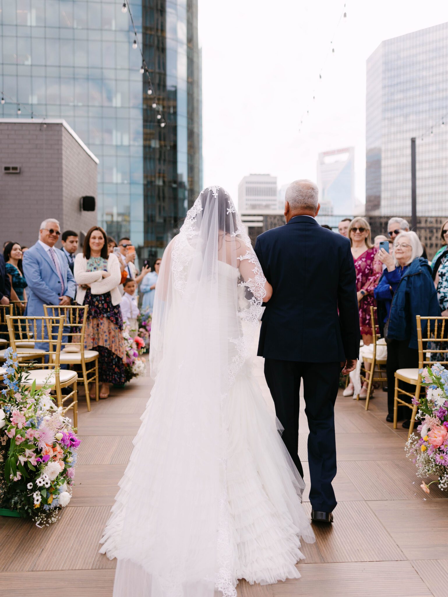 A bride in a white gown and veil walks down an outdoor aisle with an older man, surrounded by seated and standing guests. The setting is on a rooftop with city skyscrapers in the background and floral arrangements along the aisle.