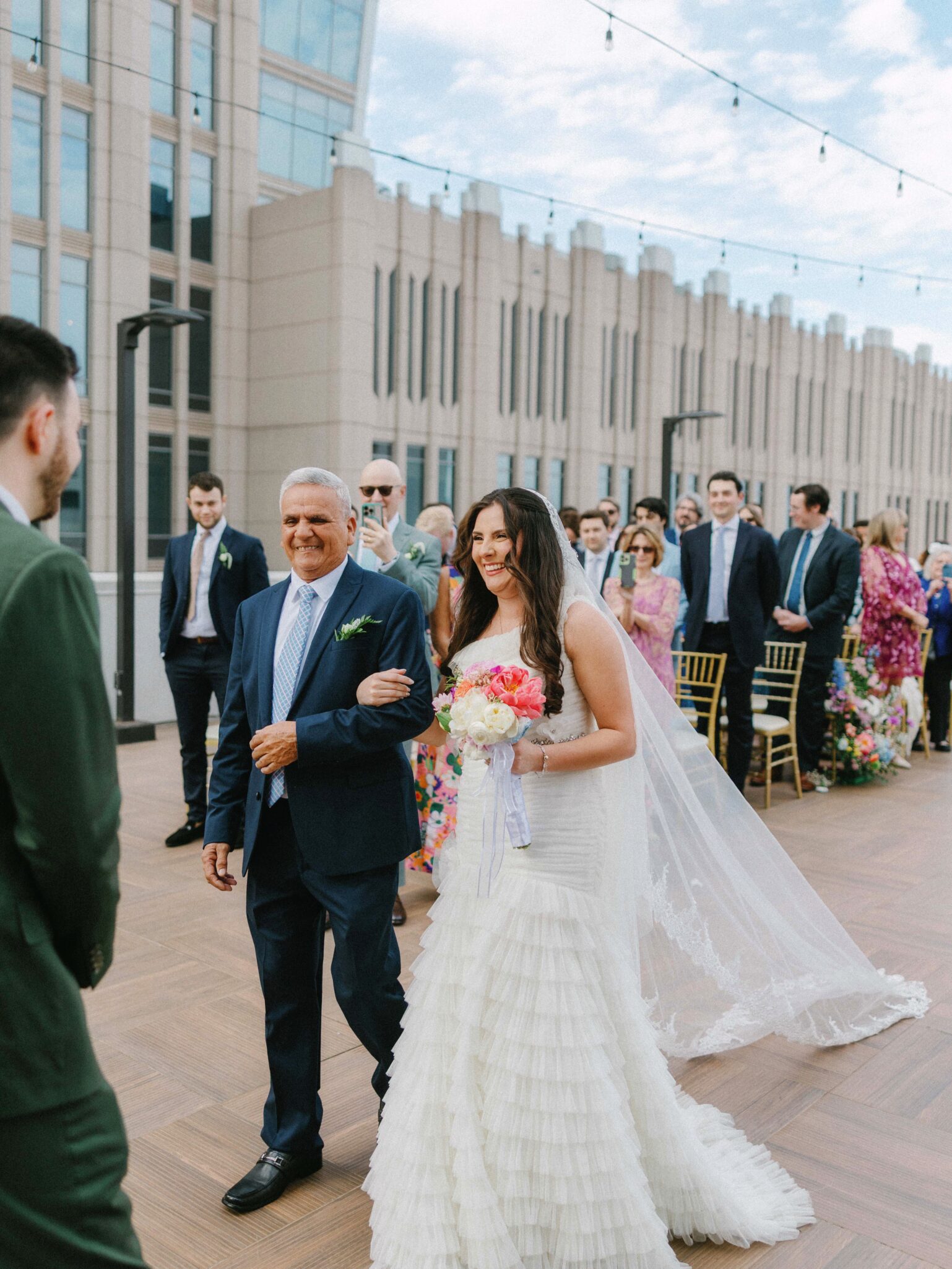 A bride in a white dress, holding a bouquet, walks down the aisle outdoors arm-in-arm with an older man in a suit. Guests watch, seated and standing, with a modern building in the background.