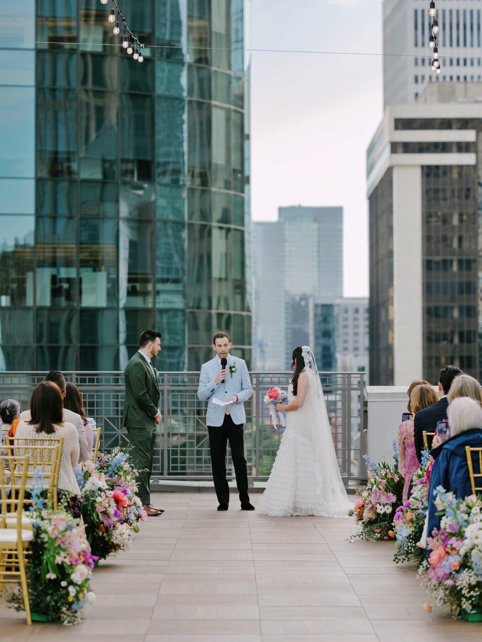 A couple stands at an outdoor Double Tree Hilton Charlotte City Center wedding ceremony, surrounded by colorful flowers and guests, with tall city buildings in the background as the officiant speaks while the bride and groom face each other.