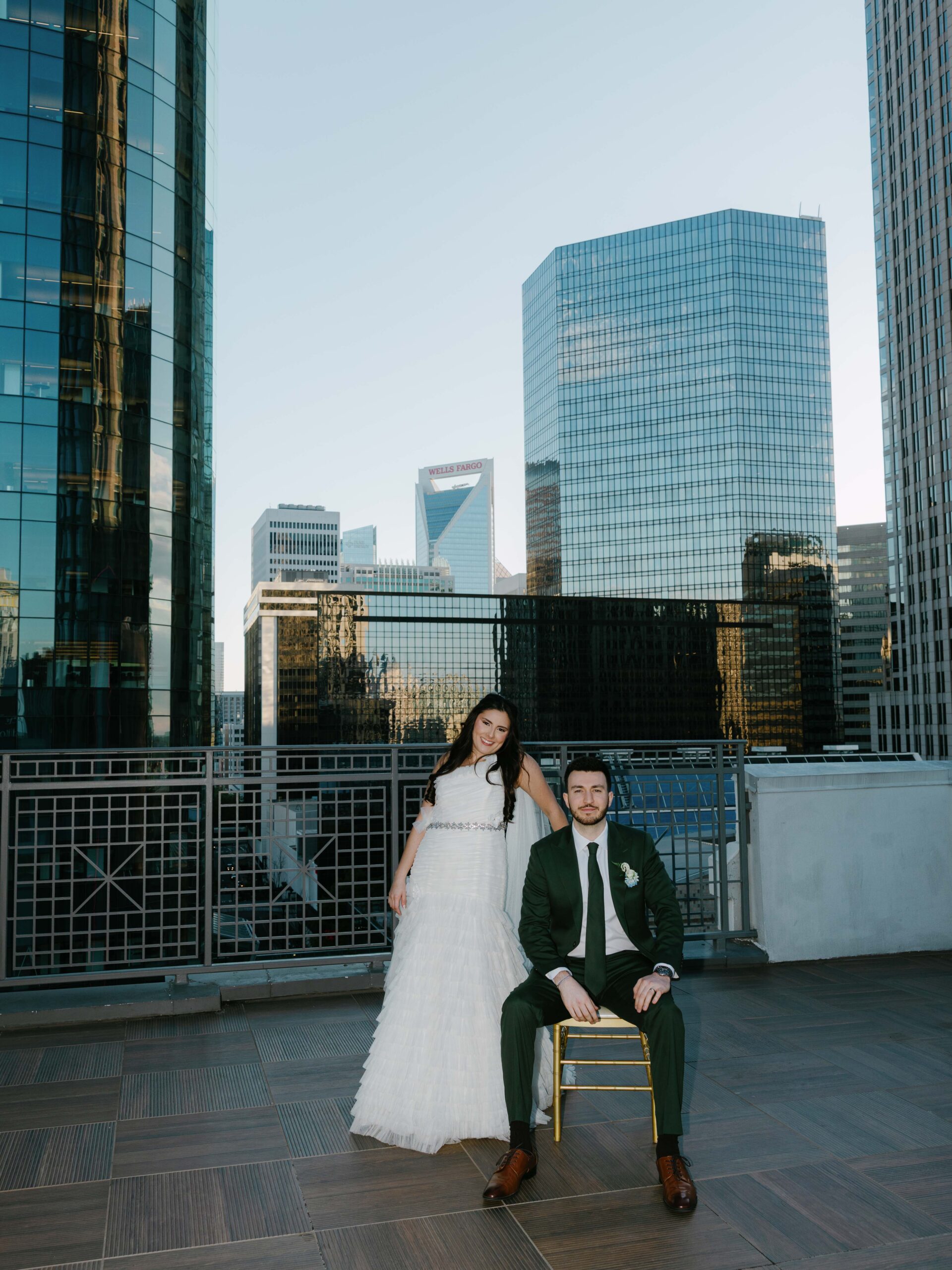 A bride in a white dress stands beside a seated groom in a dark green suit on a rooftop with modern city skyscrapers in the background.