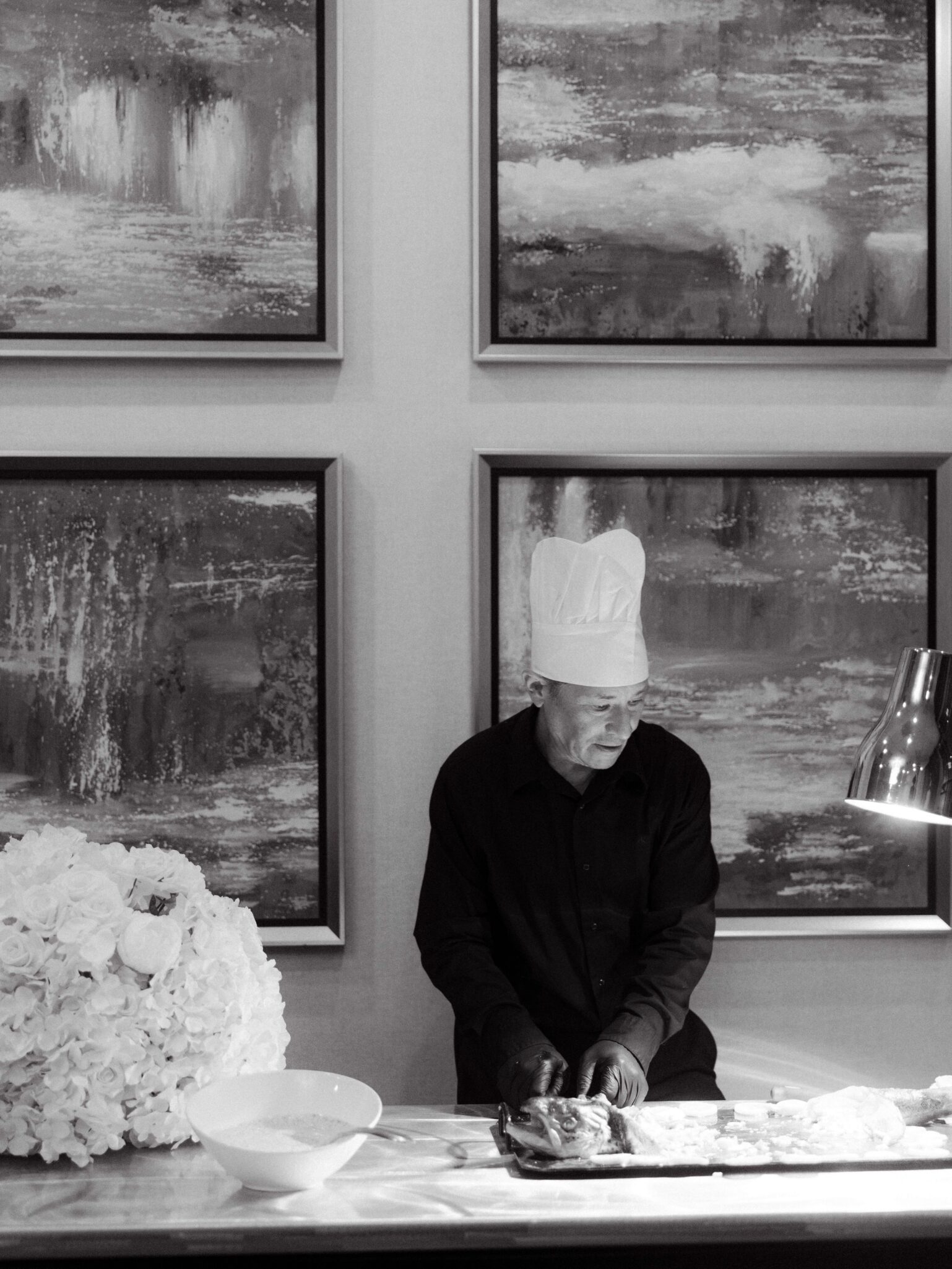 A chef in a tall white hat prepares food at a counter, with a large bouquet of flowers and a bowl nearby, during a Double Tree Hilton Charlotte City Center Wedding. Abstract paintings hang on the wall behind him. The image is in black and white.