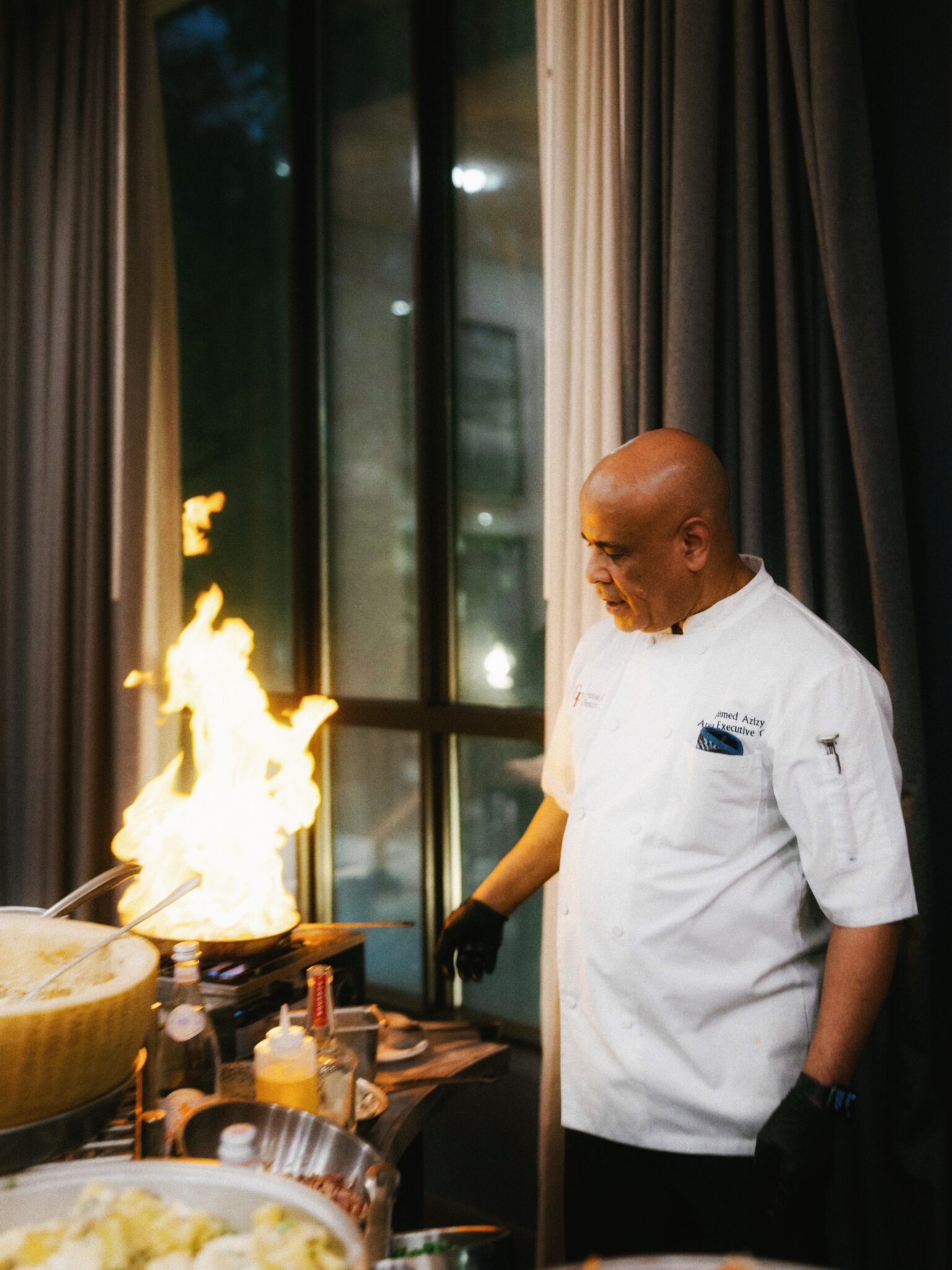 A chef wearing a white uniform and black gloves stands indoors near a flaming pan on a stovetop, surrounded by food ingredients and cooking supplies. Large windows and curtains are in the background.
