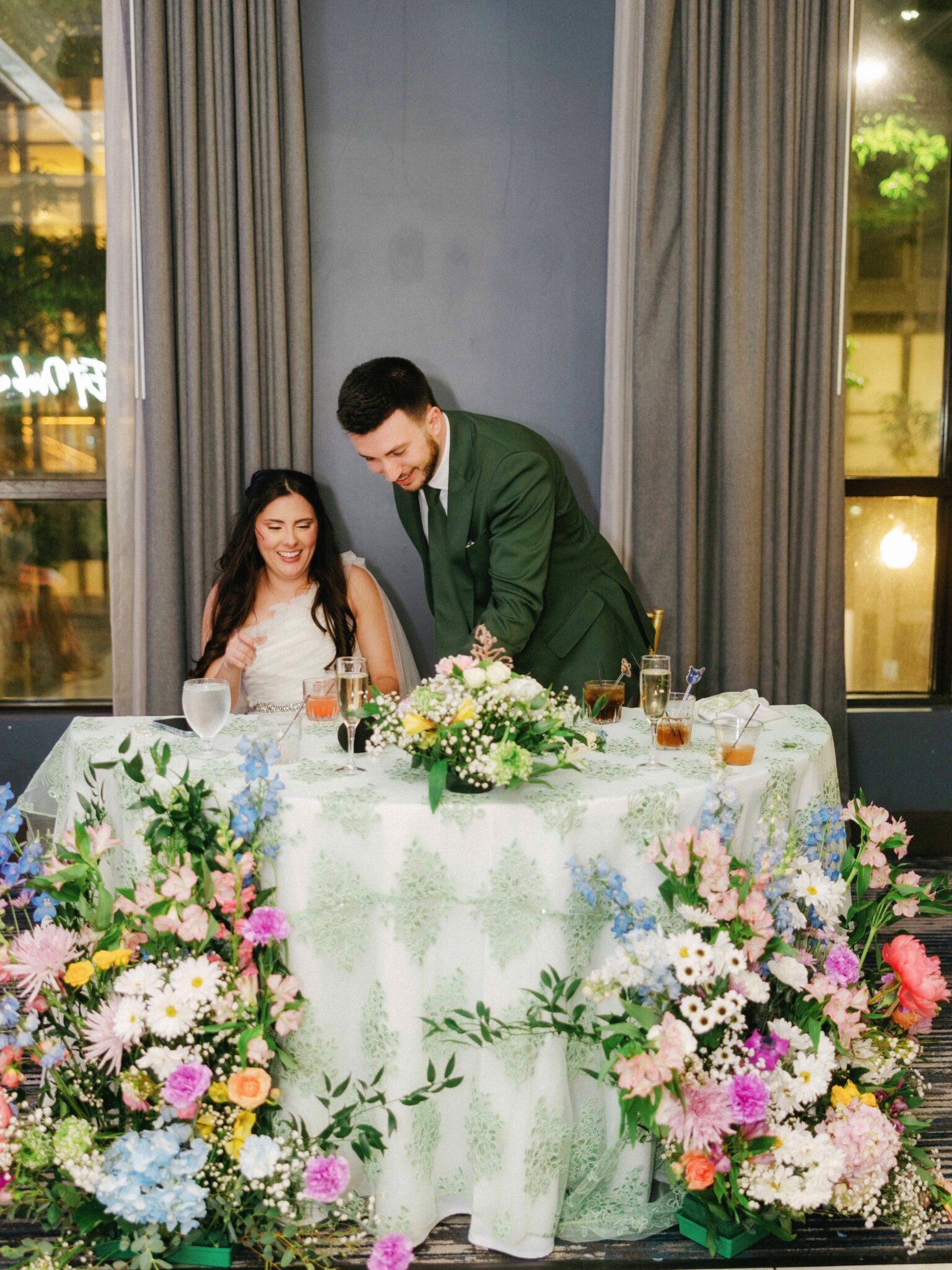 A bride and groom sit at a table adorned with pastel flowers and greenery. The groom, wearing a green suit, leans over to speak to the smiling bride. Large floral arrangements cover the front of the table.