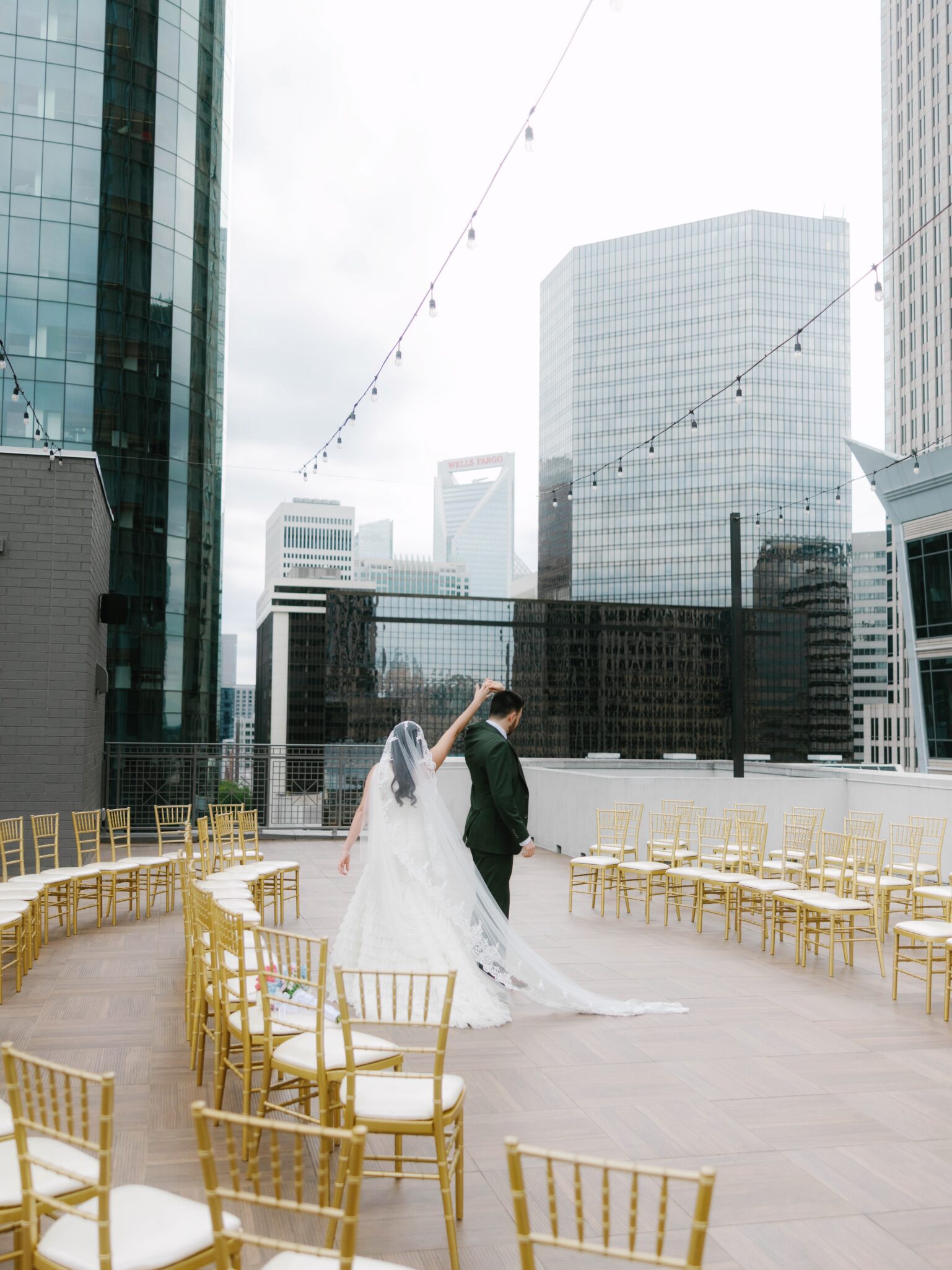 A bride and groom dance together on a rooftop at the Double Tree Hilton Charlotte City Center, surrounded by empty gold chairs, with tall city skyscrapers in the background and string lights overhead.