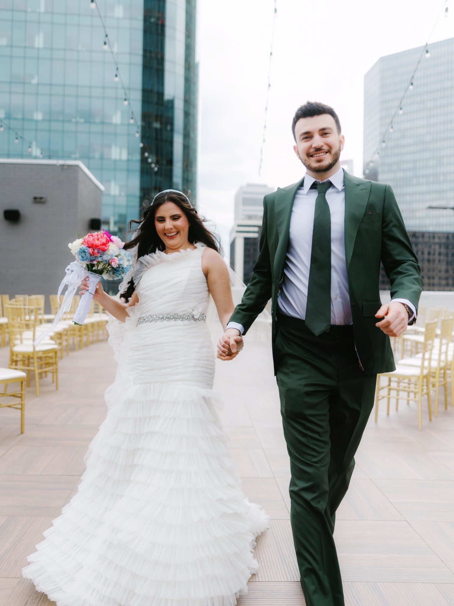 A newlywed couple holding hands and smiling, walking outdoors on a rooftop at Double Tree Hilton Charlotte City Center, decorated with string lights and gold chairs. The bride wears a white gown with bouquet; the groom sports a dark green suit and tie.