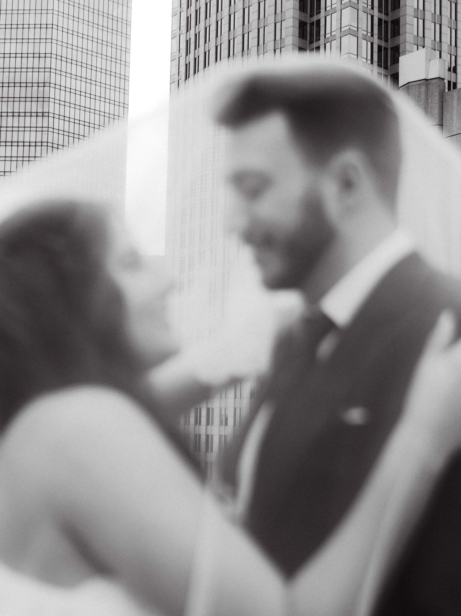 A black-and-white, softly blurred photo of a couple embracing each other in wedding attire, with tall modern buildings in the background.