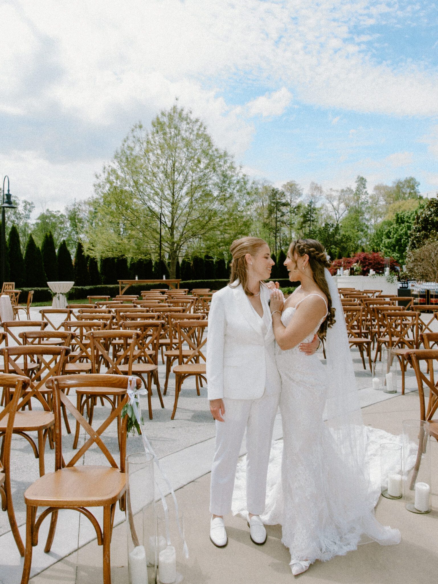 Two brides stand facing each other, holding hands and smiling, in an outdoor wedding setting with empty wooden chairs and green trees in the background under a partly cloudy sky.