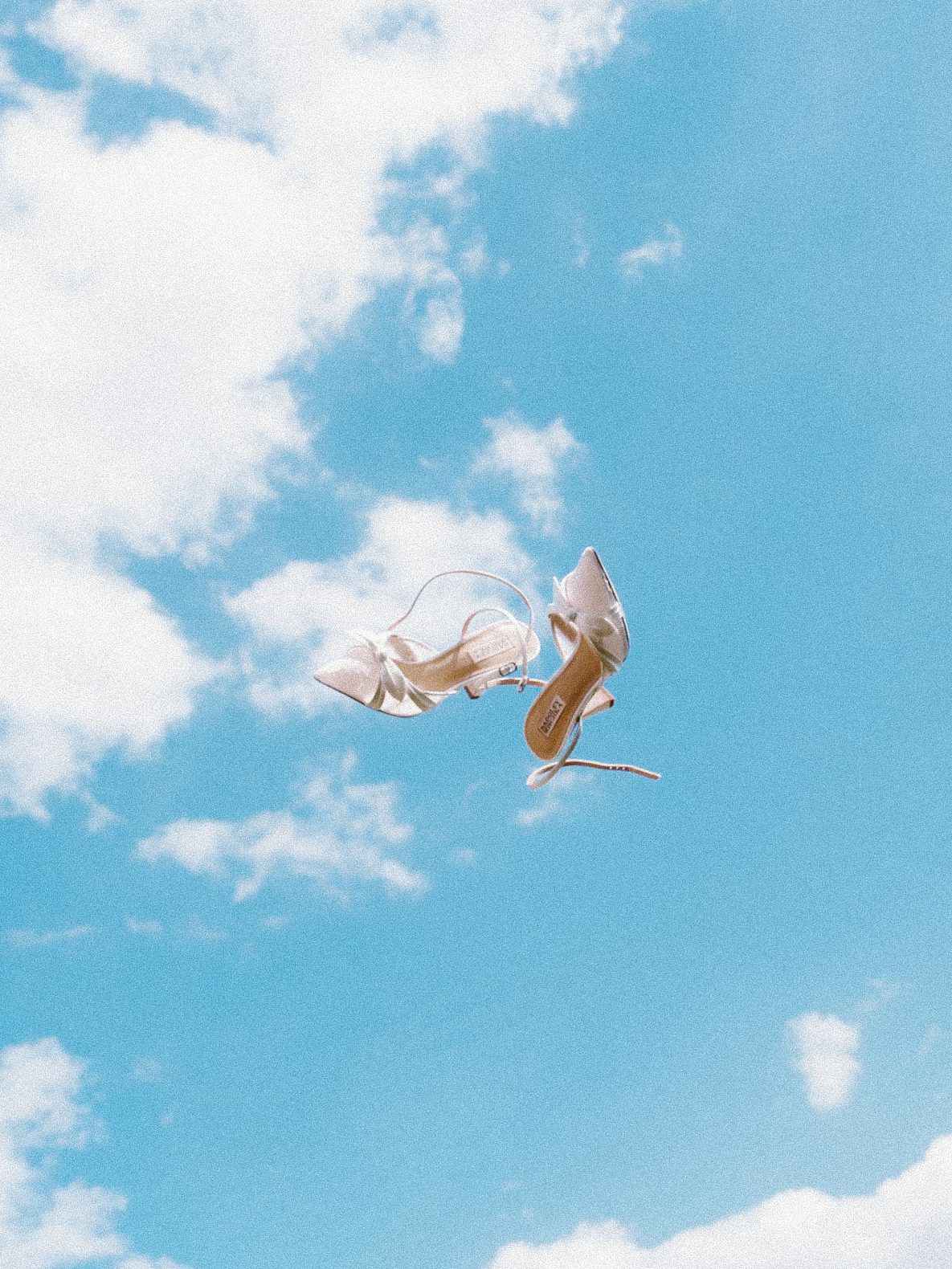A pair of white high-heeled shoes appear to be floating or falling against a bright blue Chestnut Ridge sky scattered with clouds.